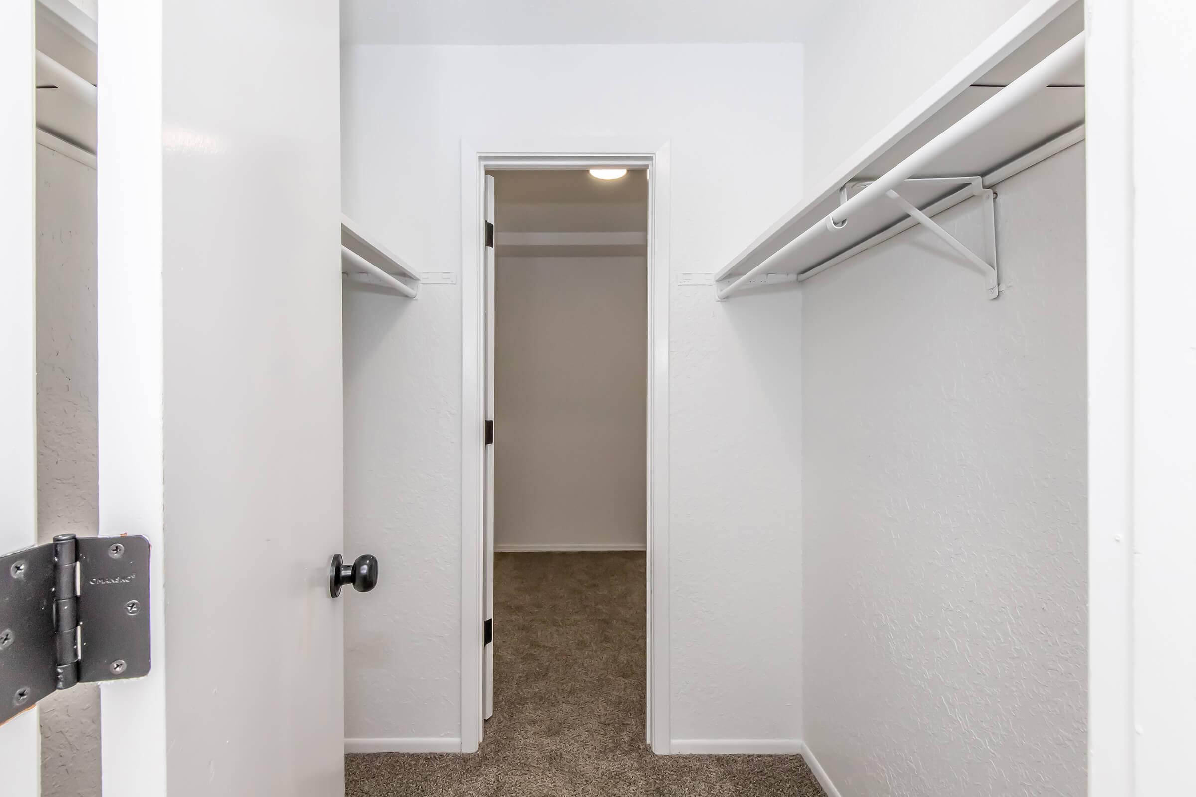 A view of a spacious closet with two hanging rods on either side and a door leading to another room. The walls are painted white, and the floor is covered in light brown carpet. The closet appears clean and organized, with plenty of space for storage.
