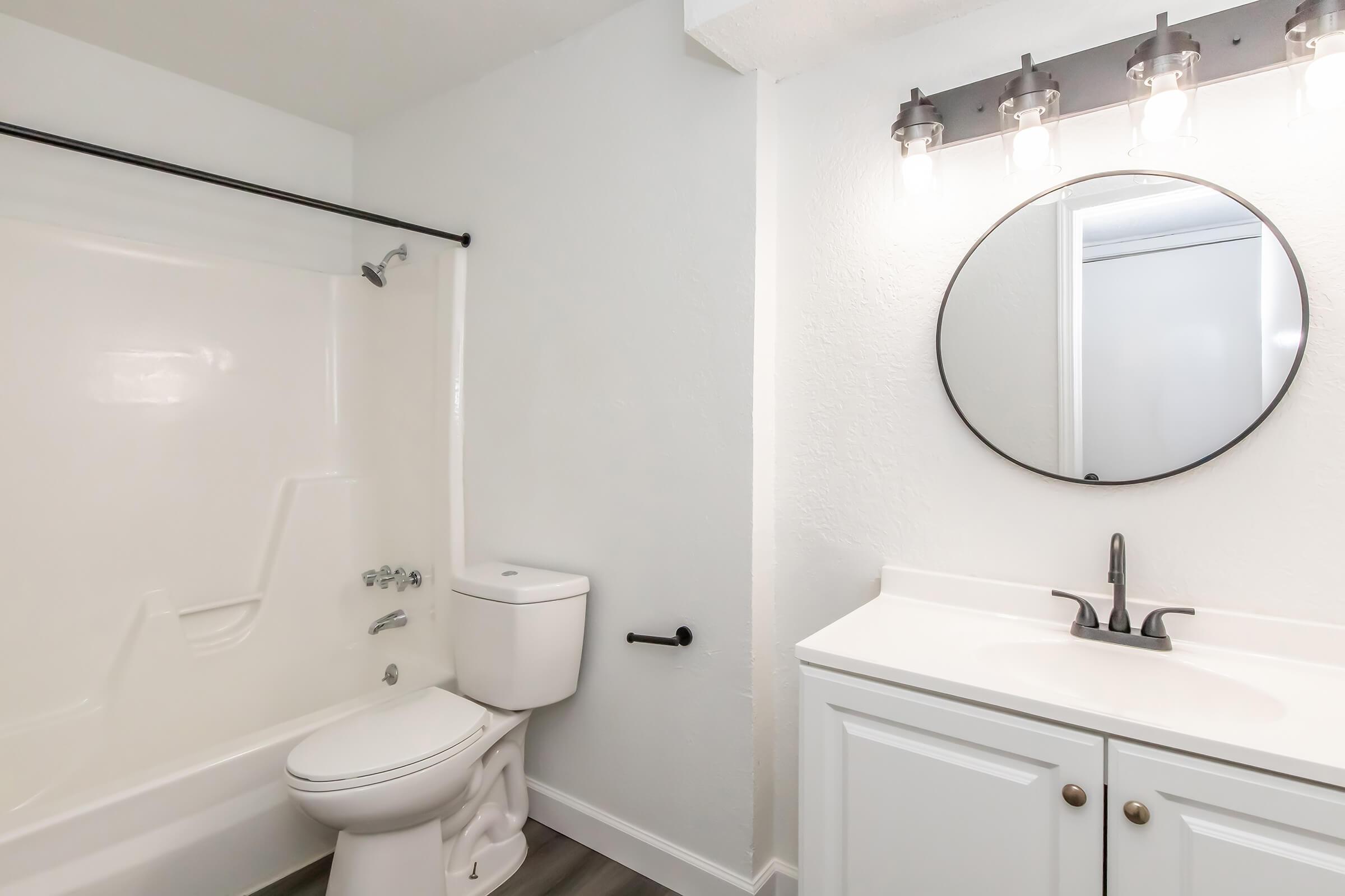 A modern bathroom featuring a white bathtub and shower combination, a toilet, and a white vanity with a sink. Above the vanity, there is a round mirror and four light fixtures. The walls are painted white, and the flooring is a light-colored wood.