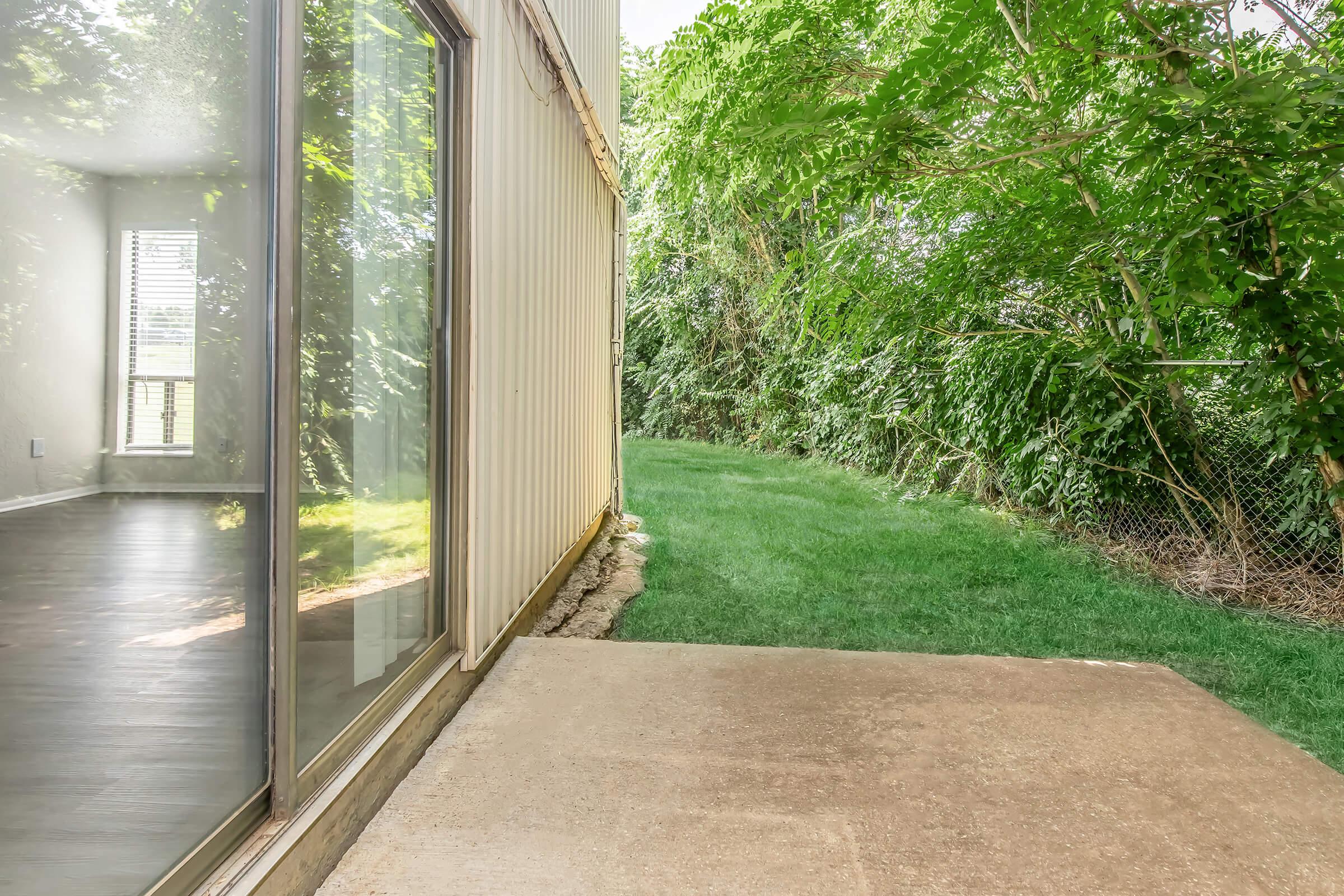 View of a grassy area beside a modern building with large glass windows reflecting the outdoors. The scene includes lush greenery and a concrete patio, suggesting a peaceful, natural setting near a residential or commercial space.
