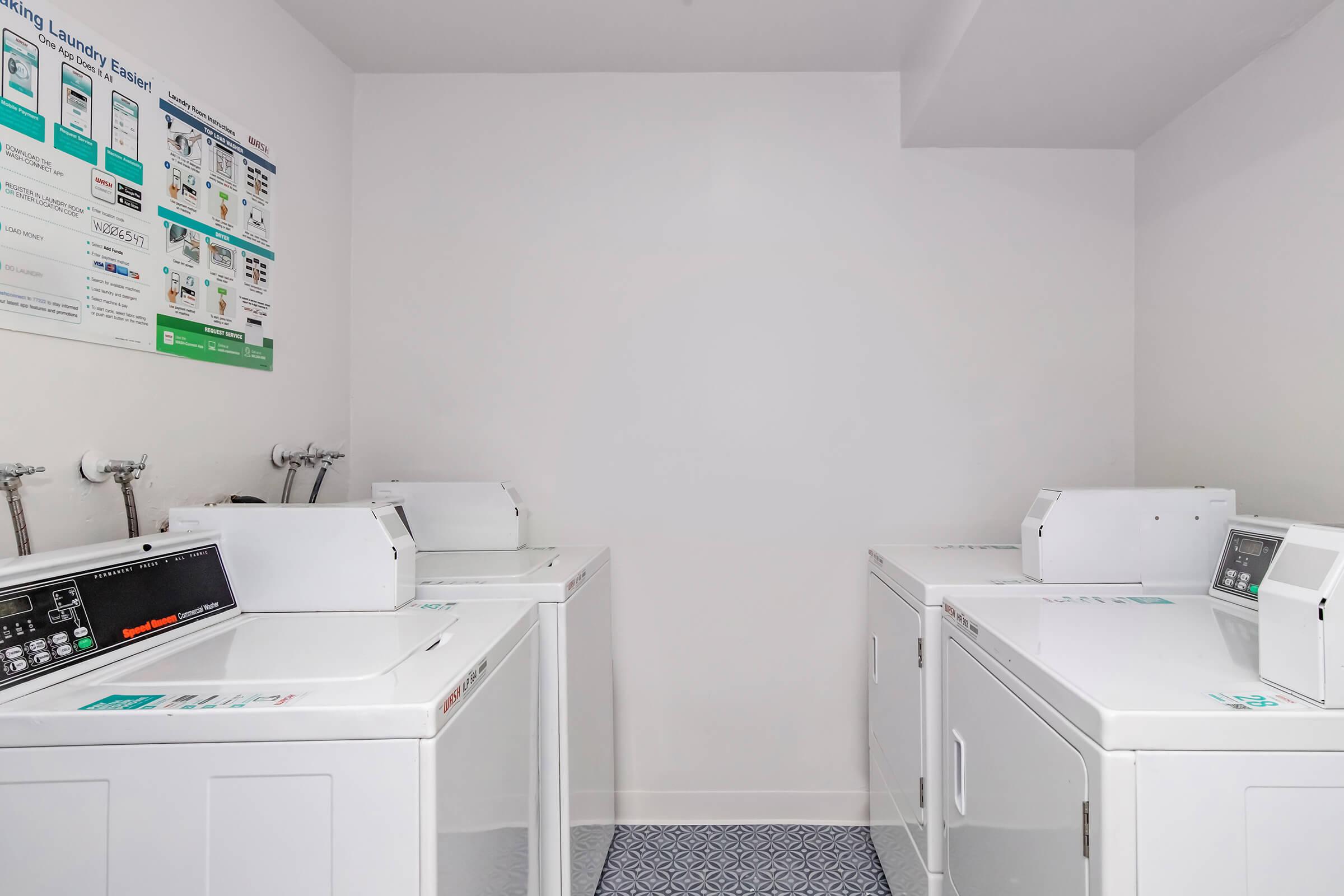 A clean, modern laundry room featuring four white washing machines and dryers aligned against a white wall. There are informational posters about laundry services on the wall, and the floor is covered with a blue patterned tile. The space is bright and well-lit.