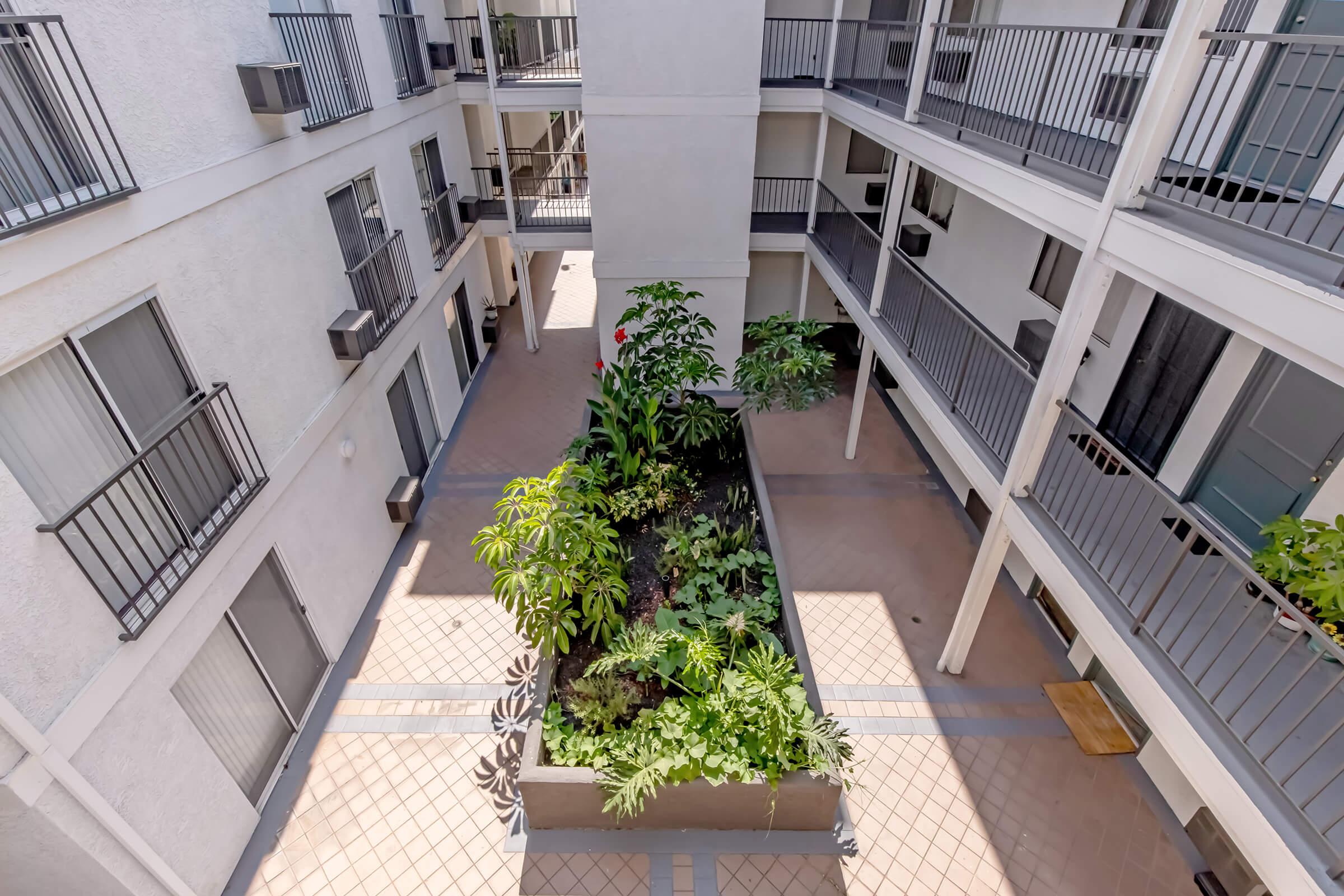A view from above of a courtyarded space featuring lush green plants and flowers in a central planter, surrounded by multiple floors of an apartment building. The area is paved with tile and has balconies on either side, creating a serene outdoor environment.