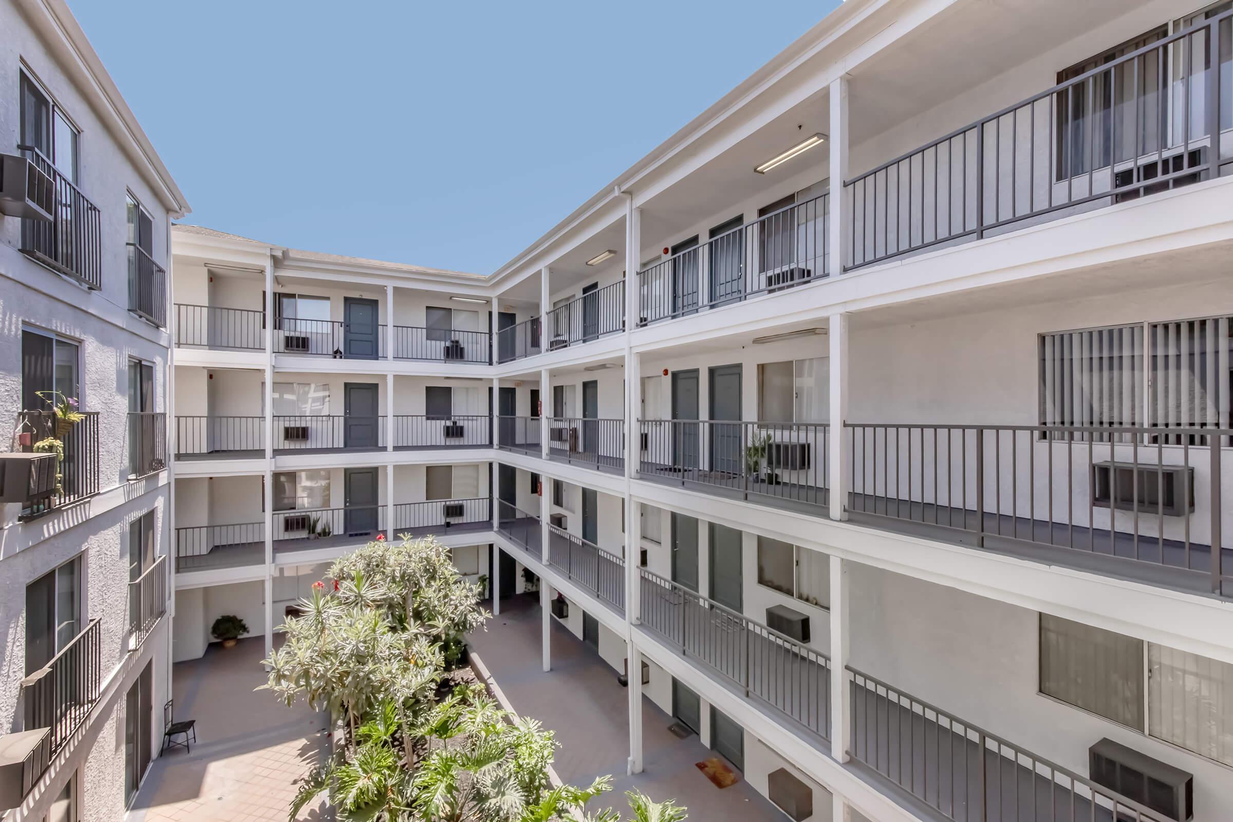 View of a multi-story apartment complex with a central courtyard. The building features multiple balconies and air conditioning units. Lush greenery is visible in the courtyard, enhancing the outdoor space. The sky above is clear and blue.