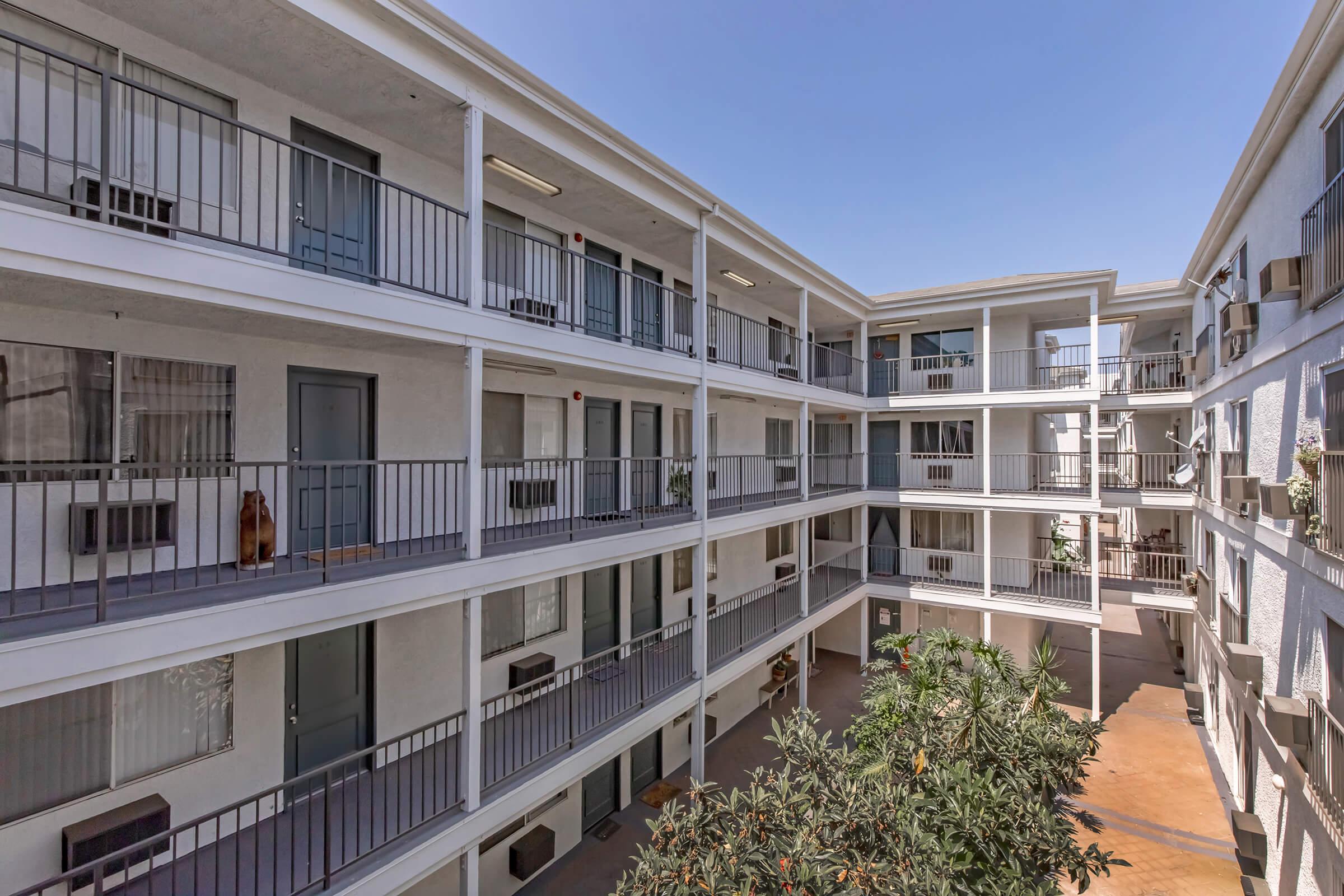A view of a modern apartment complex showcasing multiple floors with balconies. The courtyard features greenery and is surrounded by the building's architecture, capturing a bright blue sky above.