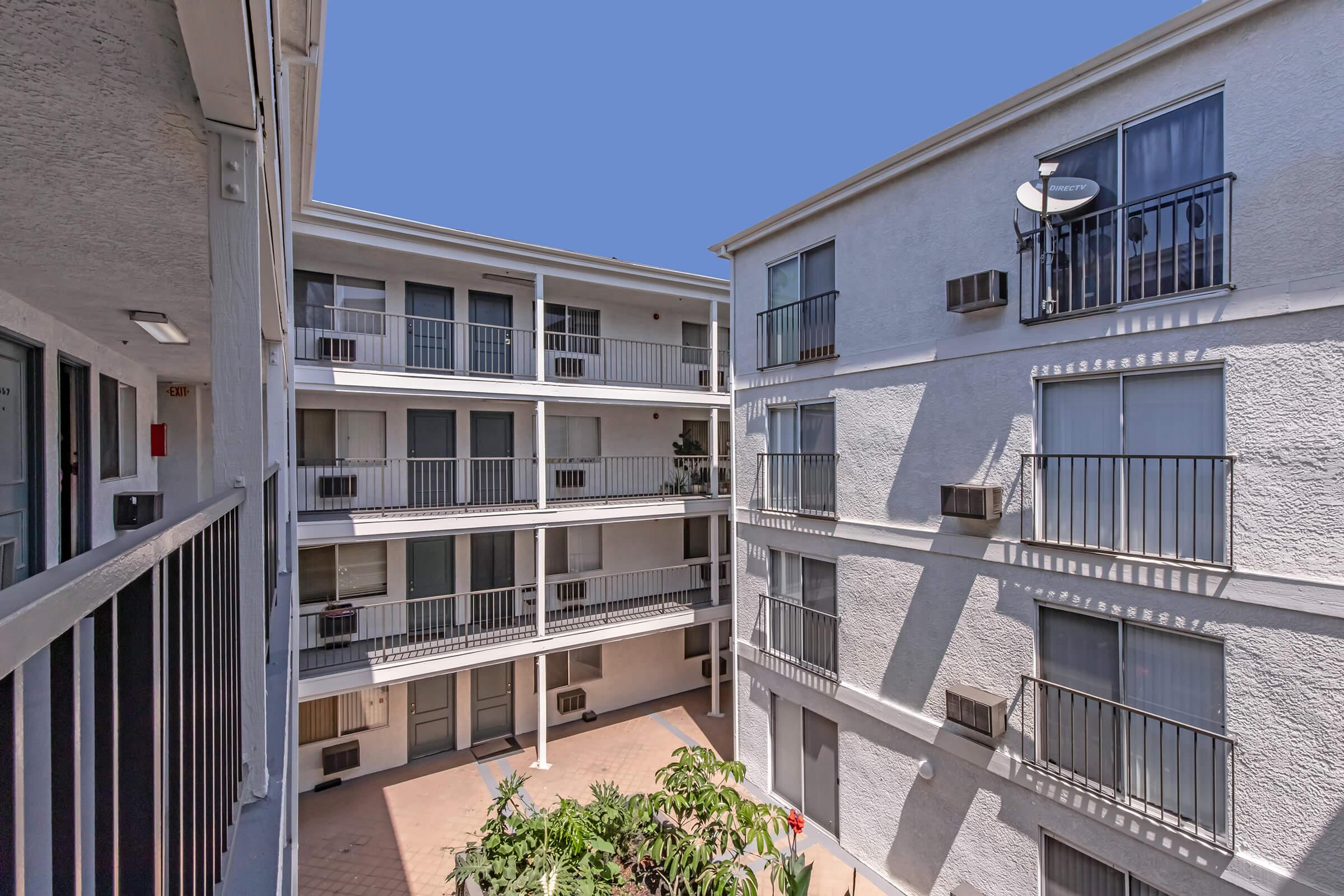 A view of a courtyard area between two multi-story buildings, featuring balconies, air conditioning units, and greenery in the foreground. The sky is clear and blue, providing a bright backdrop to the architecture. The design showcases a typical apartment complex layout.