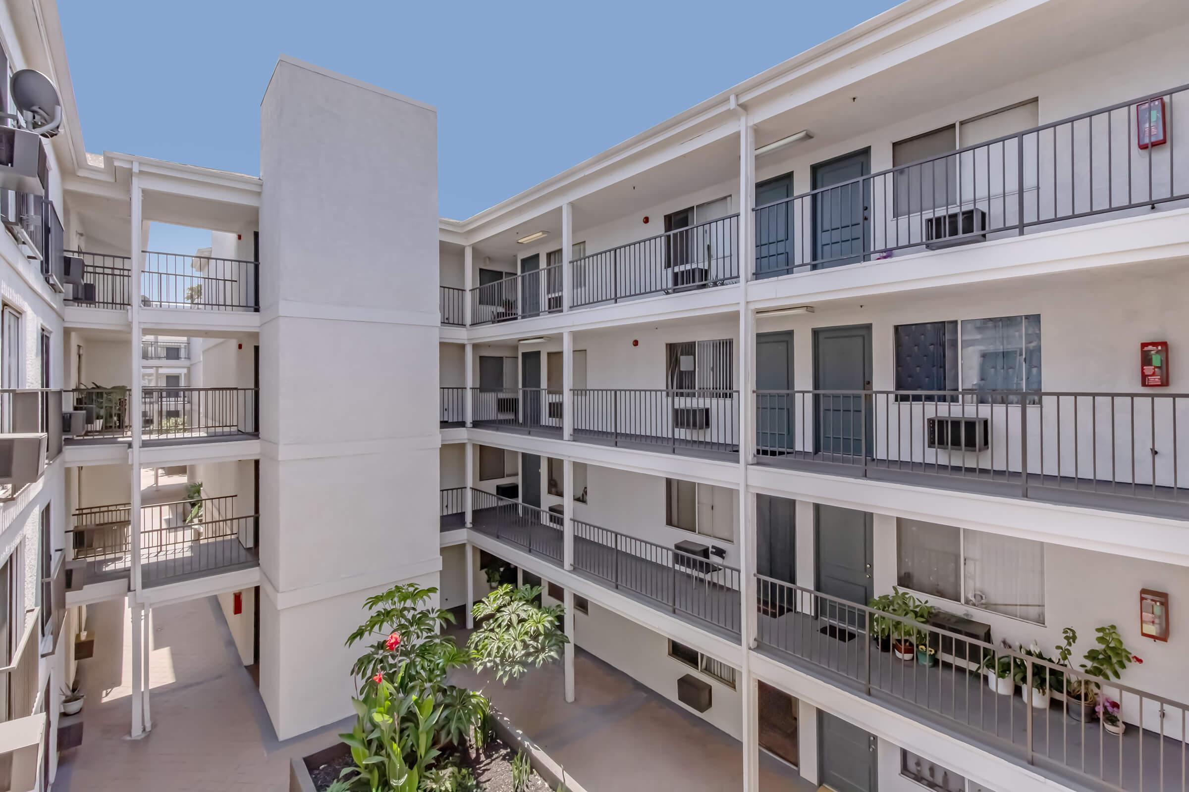 View of a multi-level apartment complex courtyard. The building features multiple balconies, plants, and air conditioning units. The sky is clear, creating a bright atmosphere in the open space.