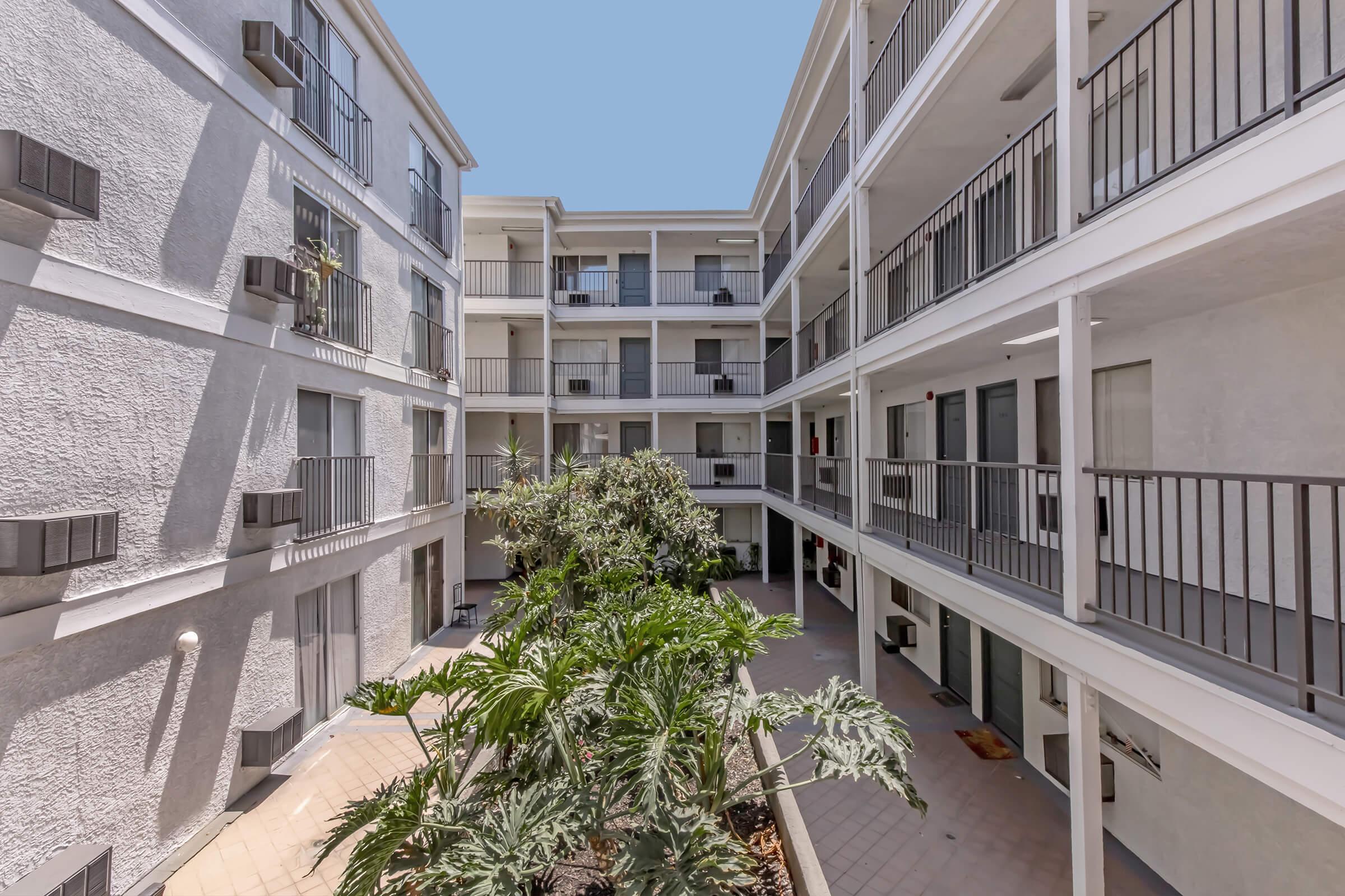 A view of a multi-story apartment building courtyard, featuring balconies on each floor, with plants and greenery in the center. The exterior walls are light-colored, and air conditioning units are visible. The sky is clear, creating a bright ambiance in the shared space.
