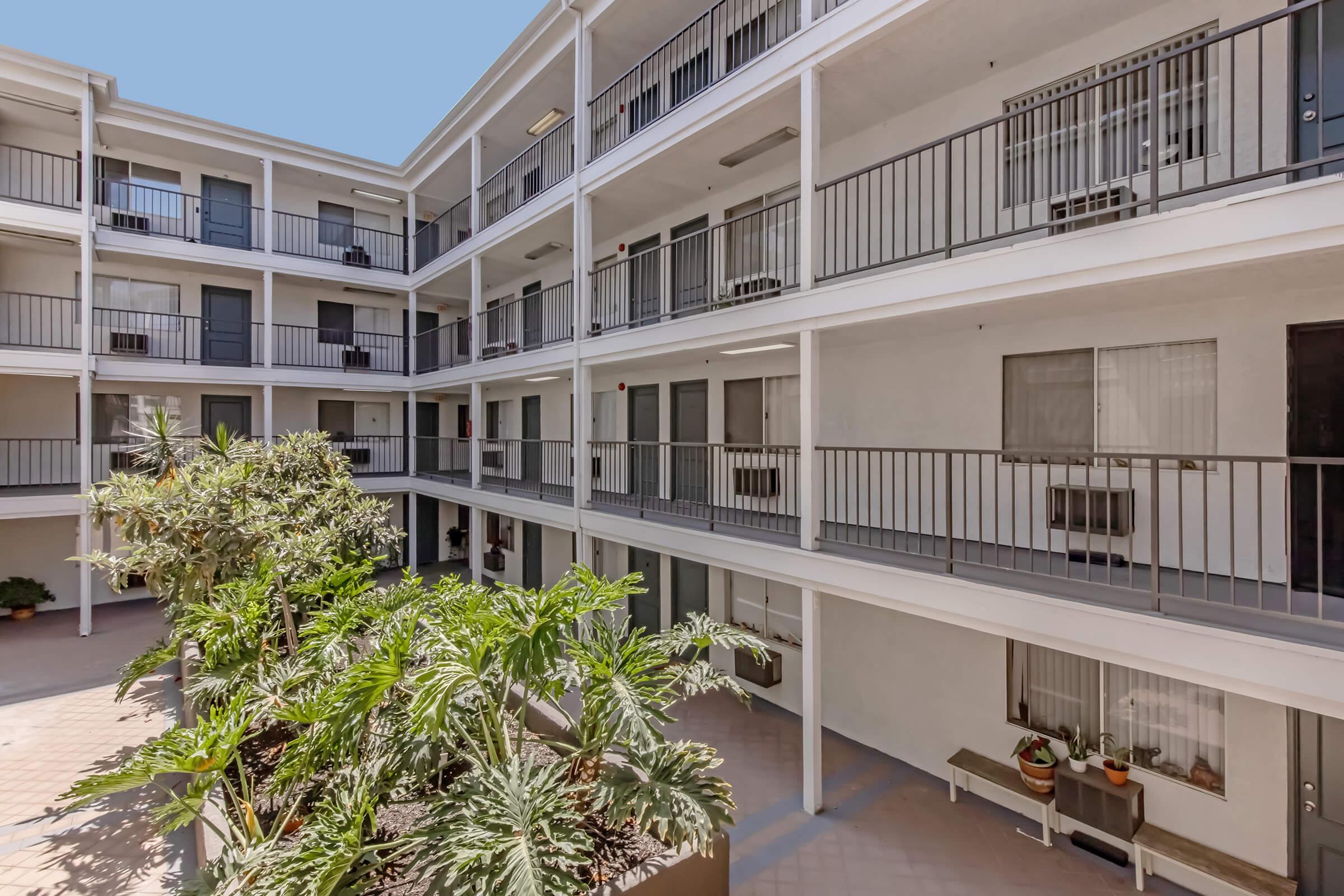 A view of an interior courtyard of a multi-story residential building. The courtyard features lush green plants, with balconies visible on both sides. Each balcony has sliding glass doors and railings, creating a bright and open atmosphere. The sky is clear blue above the structure.
