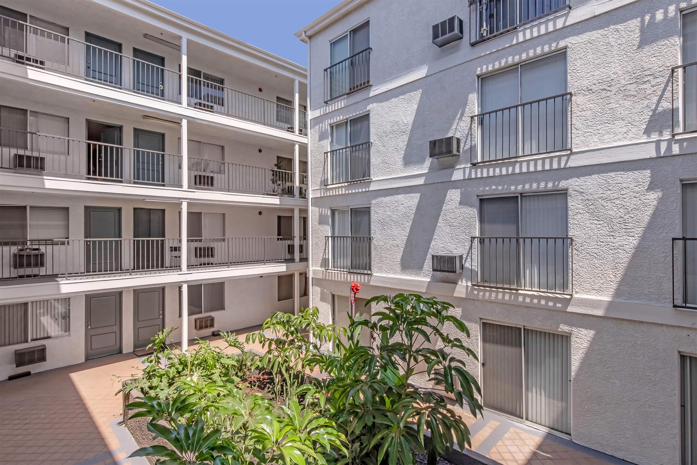 A view of a courtyard surrounded by a two-story apartment building. The courtyard features several green plants and shrubs, with a few balconies visible on the building's exterior. Sunlight illuminates the area, creating a bright and inviting atmosphere.