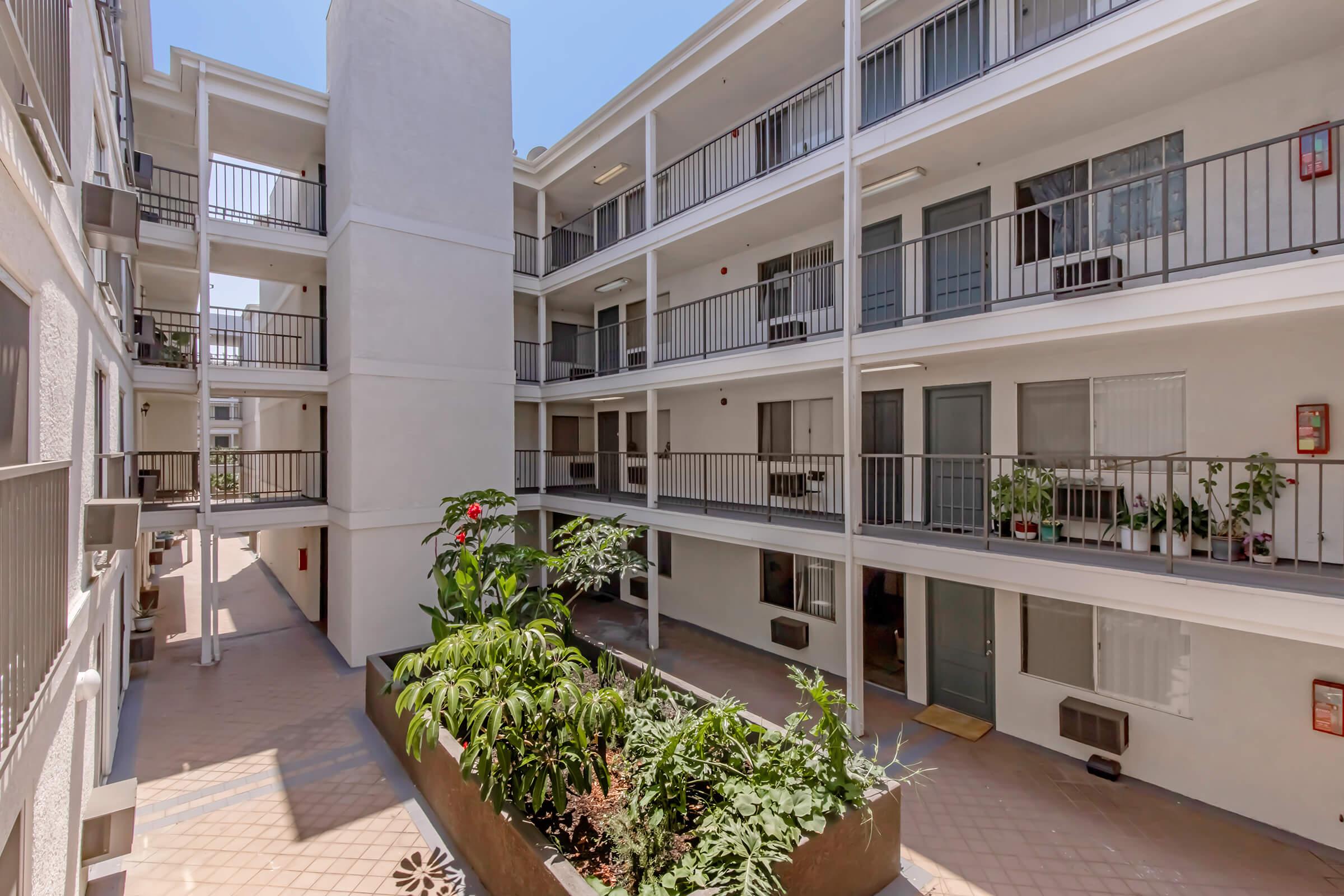 A view of a multi-story apartment building courtyard featuring several balconies, a central planter with greenery, and sunlight streaming in, creating a bright atmosphere. The surrounding architecture is modern, with multiple doors and windows visible on the upper levels.