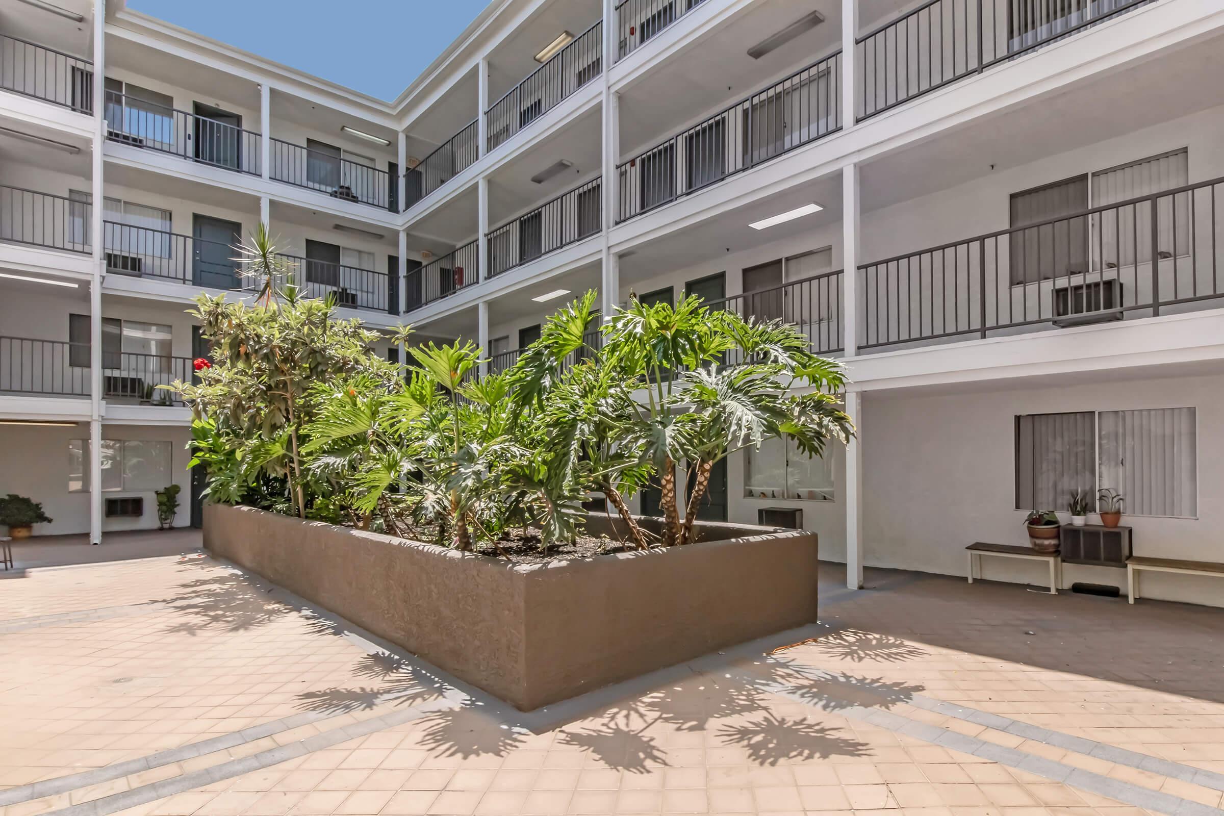 A tranquil courtyard featuring lush greenery and potted plants surrounded by a modern building with multiple balconies. The area is brightly lit by natural sunlight, creating a serene atmosphere suitable for relaxation.