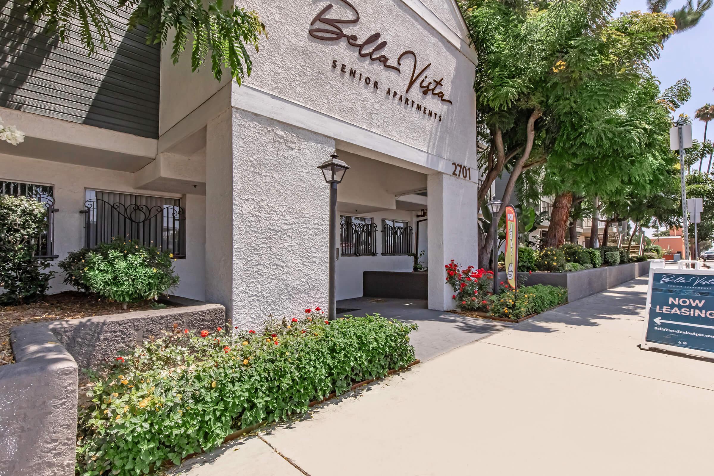 Exterior view of Bella Vita Senior Apartments, featuring a welcoming entrance with a landscaped pathway, colorful flowering plants, and decorative lampposts. A leasing sign is visible nearby, indicating that apartments are currently available for rent. Surrounding greenery enhances the inviting atmosphere.