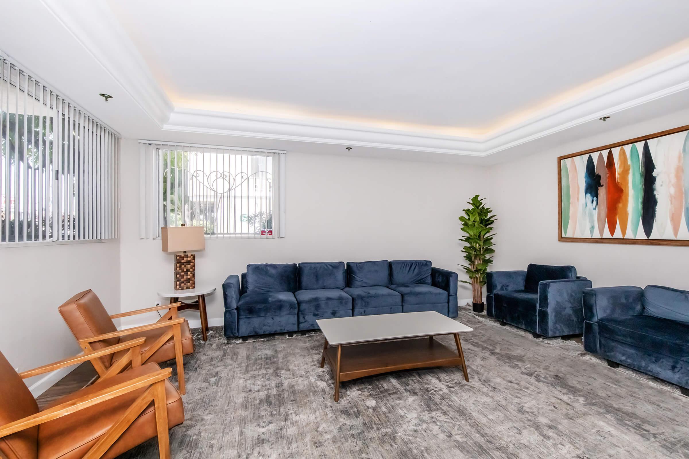 A modern, minimalist living room featuring a blue velvet sectional sofa, matching chairs, a wooden coffee table, and a potted plant. Soft lighting highlights the space, with large windows and blinds allowing natural light in. A colorful abstract art piece decorates one wall, adding a vibrant touch.