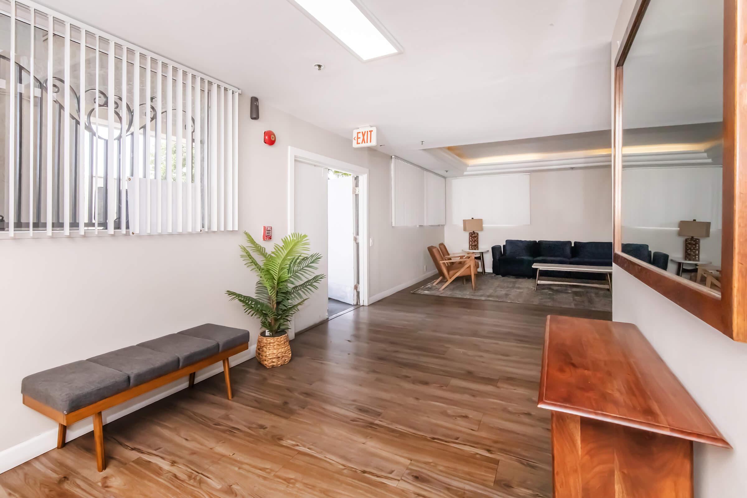 A bright hallway with wooden flooring featuring a bench, a potted plant, and a mirror. There is an exit door visible on the left, and a cozy seating area at the end of the hall with a dark-colored couch and a coffee table. Natural light illuminates the space through the windowed wall.