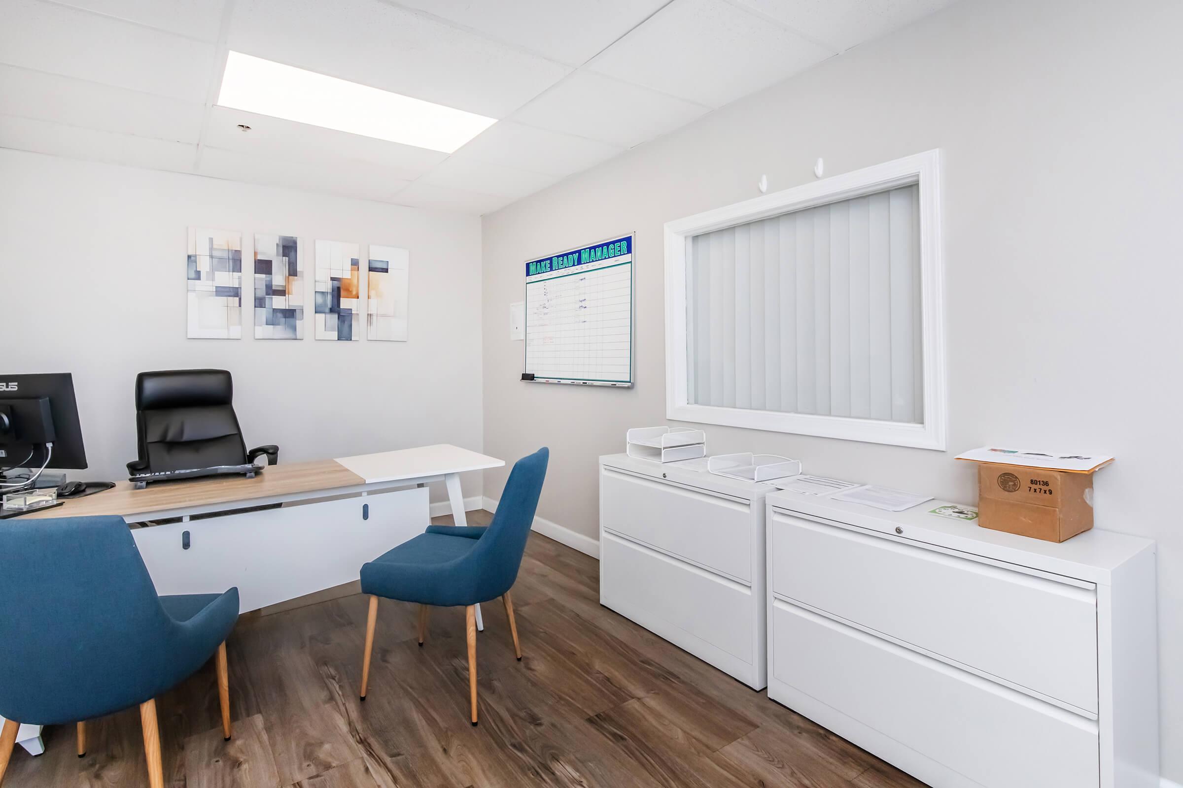 A modern office interior featuring a white desk with a computer and a black chair. There are two blue chairs in front of the desk, a wall-mounted calendar, and a window with blinds. The walls are painted light gray, and abstract artwork is displayed on one wall. Drawer cabinets are positioned against another wall.