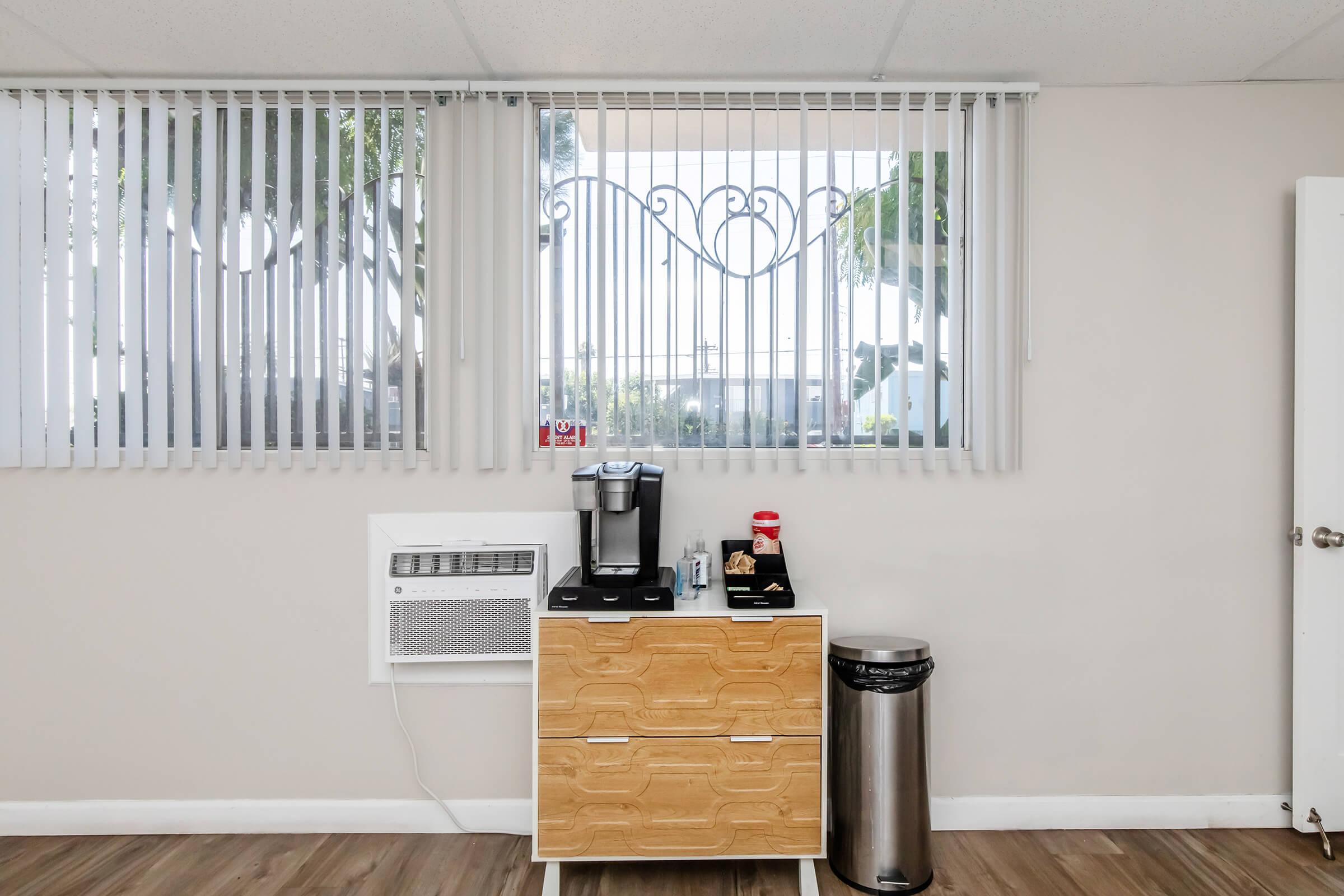 A light-colored room featuring a window with vertical blinds. Below the window, there is a coffee maker on a wooden cabinet with a few kitchen supplies. To the right, there's a trash can and an air conditioning unit beneath the window. The walls are neutral, creating a clean and modern appearance.