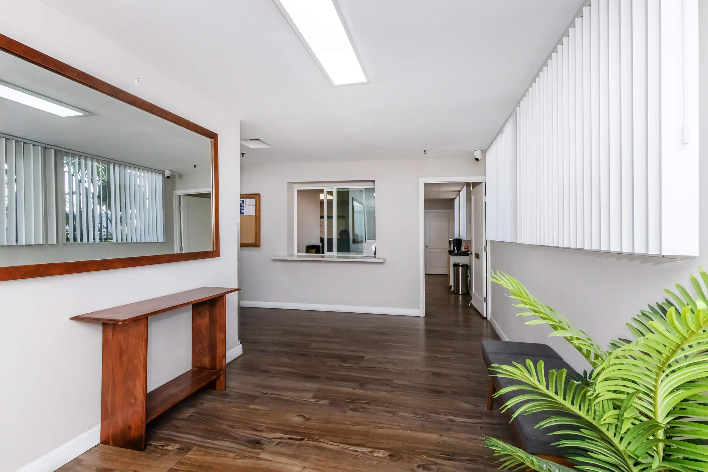 A spacious interior hallway with wood flooring, featuring a large mirror on the left and a wooden console table. On the right, there is a reception area with a small counter behind a partition. Sunlight filters in through the window blinds, and a green plant adds a touch of nature to the scene.