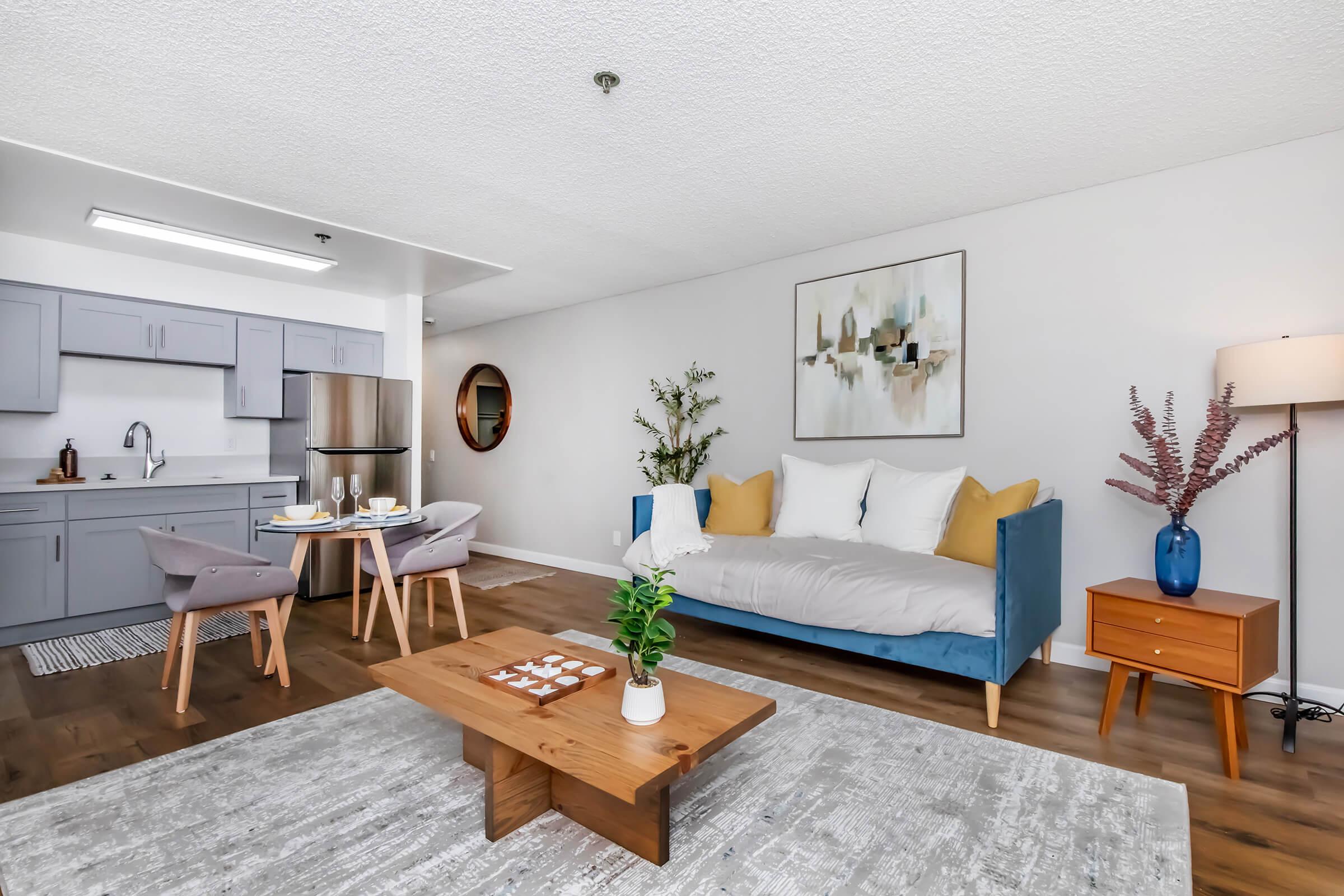A modern living room featuring a blue couch with yellow and white cushions, a wooden coffee table, and a tasteful area rug. There is a small dining set with two chairs and a kitchen in the background. A decorative plant and a floor lamp add to the inviting atmosphere, along with wall art and a round mirror.