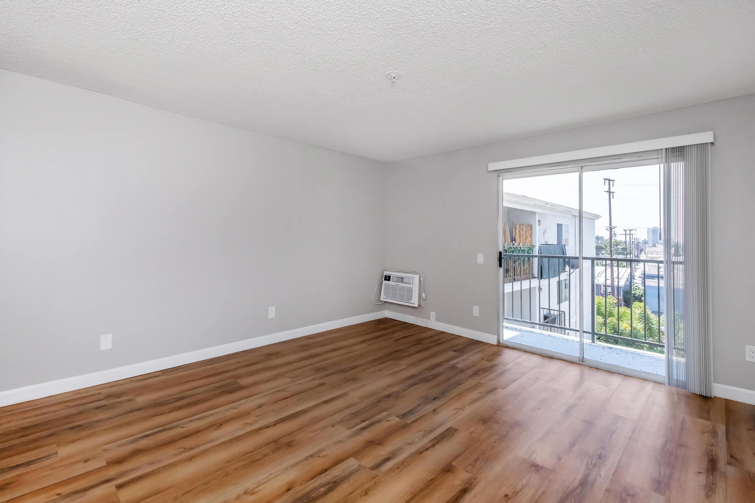 A spacious, empty room featuring light gray walls and a wooden floor. A large sliding glass door leads to a small balcony with a view. An air conditioning unit is installed on one wall. The overall ambiance is bright and minimalist, suitable for various decorative styles.