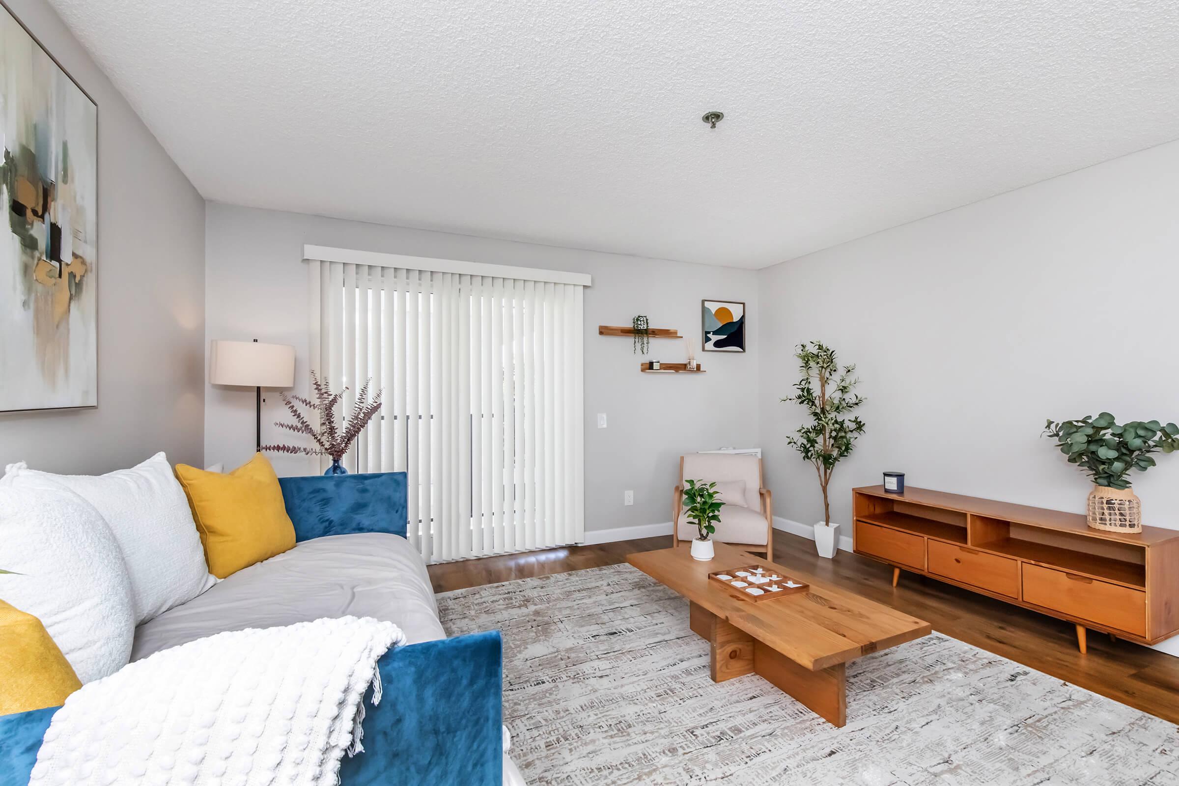 A cozy living room featuring a blue and white sofa with yellow cushions, a wooden coffee table, and a TV console. The space has light-colored walls, a textured rug, and potted plants. Natural light filters in through vertical blinds, and there are decorative items on the walls and furniture, creating a warm atmosphere.
