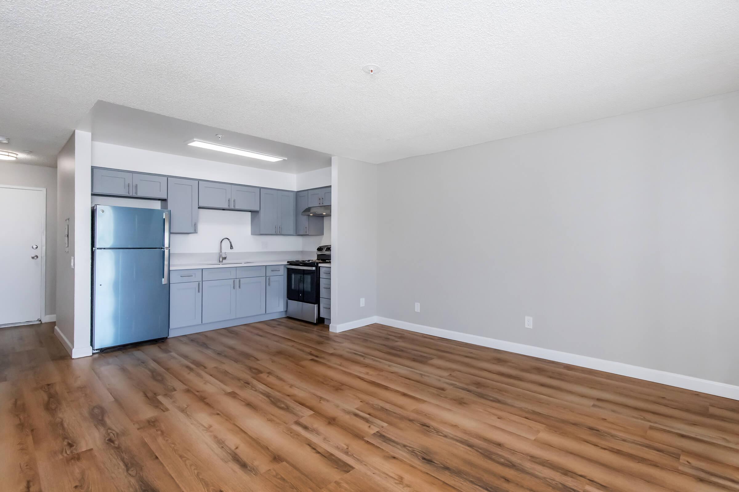 Interior view of a modern kitchen and living area featuring gray cabinets, stainless steel appliances, a refrigerator, and wooden flooring. The space is well-lit with a neutral color scheme, showcasing an open layout with ample room for furniture.