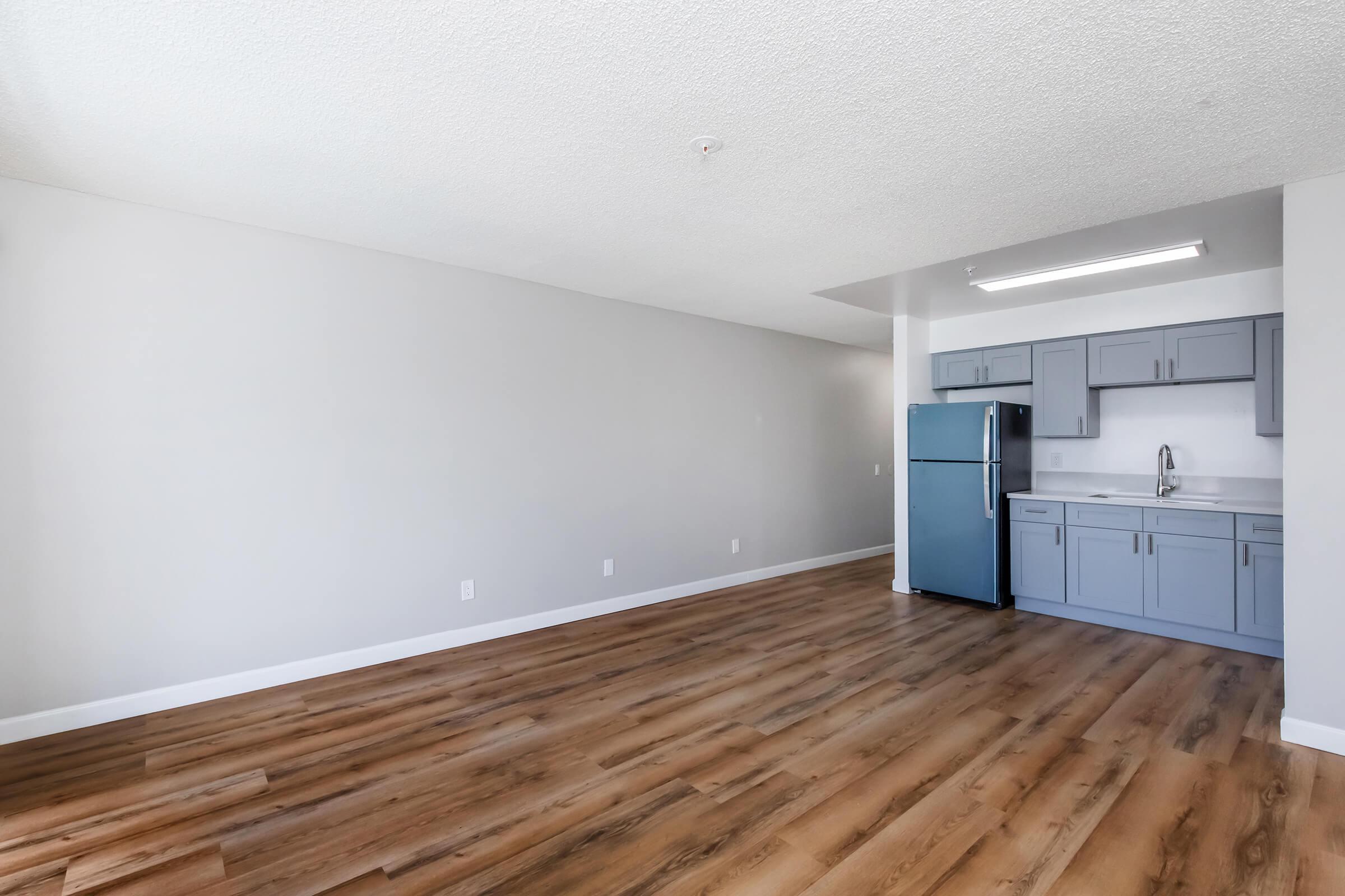 A spacious, empty apartment with light gray walls and wooden laminate flooring. The kitchen features blue cabinetry and a stainless steel refrigerator. The area is well-lit with overhead lighting and has an open layout, providing a bright and inviting atmosphere.