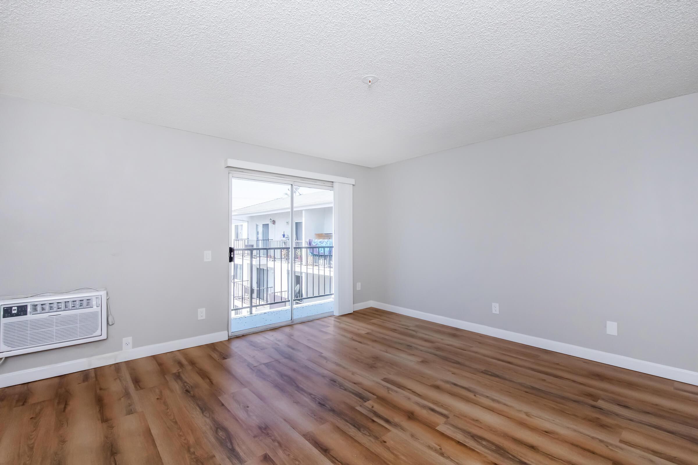 A bright, unfurnished living room featuring light gray walls and a large window with a sliding glass door that leads to a balcony. The flooring is a warm wooden laminate. An air conditioning unit is mounted on the left wall, and the space is empty, ready for furniture arrangement.