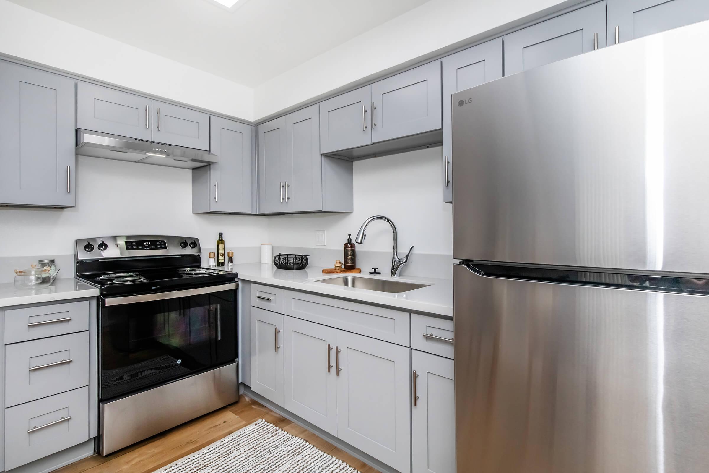 Modern kitchen featuring gray cabinets, a stainless steel refrigerator, an oven, and a sink. The countertop is white, complemented by wooden flooring and a textured rug. The space includes decorative items like bottles and a bowl, creating a welcoming and functional atmosphere.