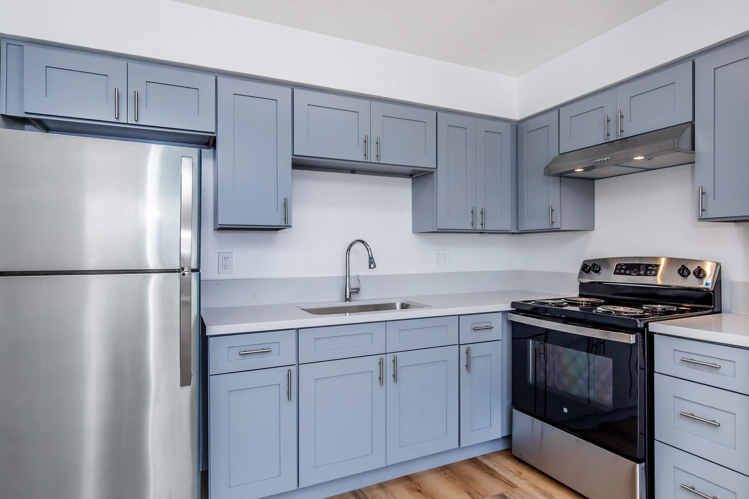 A modern kitchen featuring light blue cabinetry, a stainless steel refrigerator, a stainless steel stove, and a sink. The countertops are white, and there's hardwood flooring. The design is clean and contemporary, with ample storage and a well-lit atmosphere.