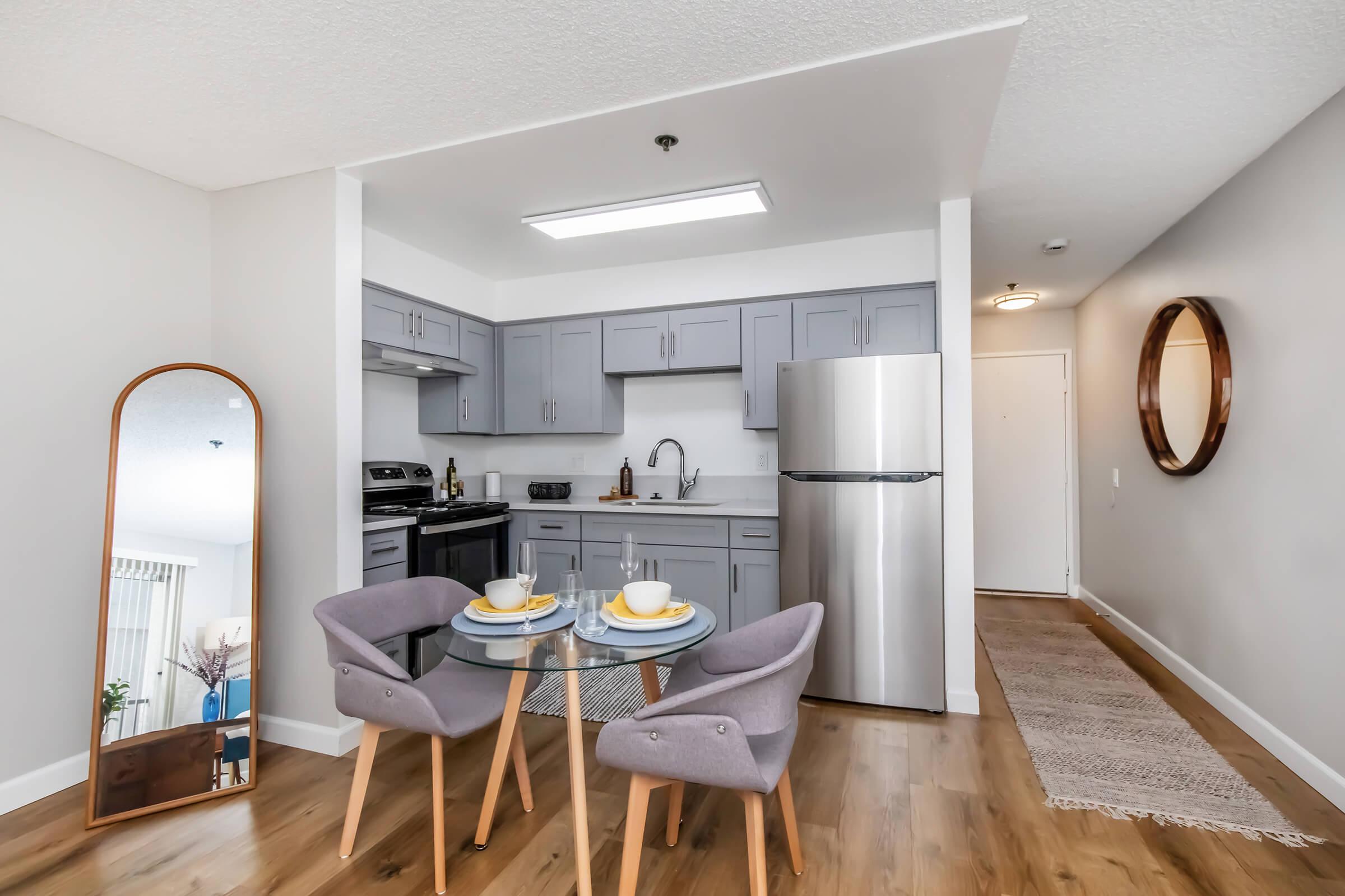 Modern kitchen and dining area featuring gray cabinetry, stainless steel appliances, and a round glass dining table set for two. The space includes a floor mirror and warm wooden flooring, with a doorway leading to a hallway. Bright, neutral walls create an inviting atmosphere.