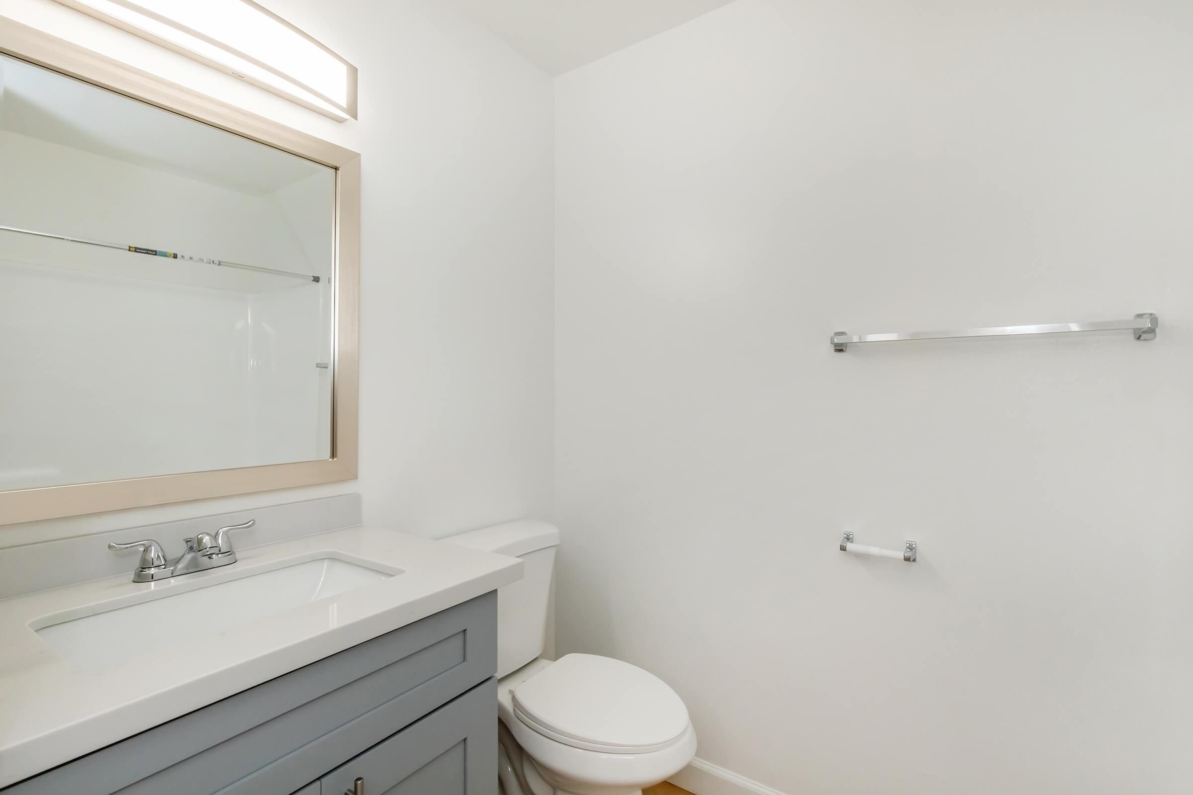 A clean and modern bathroom featuring a light-colored wall, a rectangular mirror with a simple frame above a white sink, a gray cabinet underneath, a white toilet, and a towel bar mounted on the wall with minimal decor. The space has a bright and airy feel.