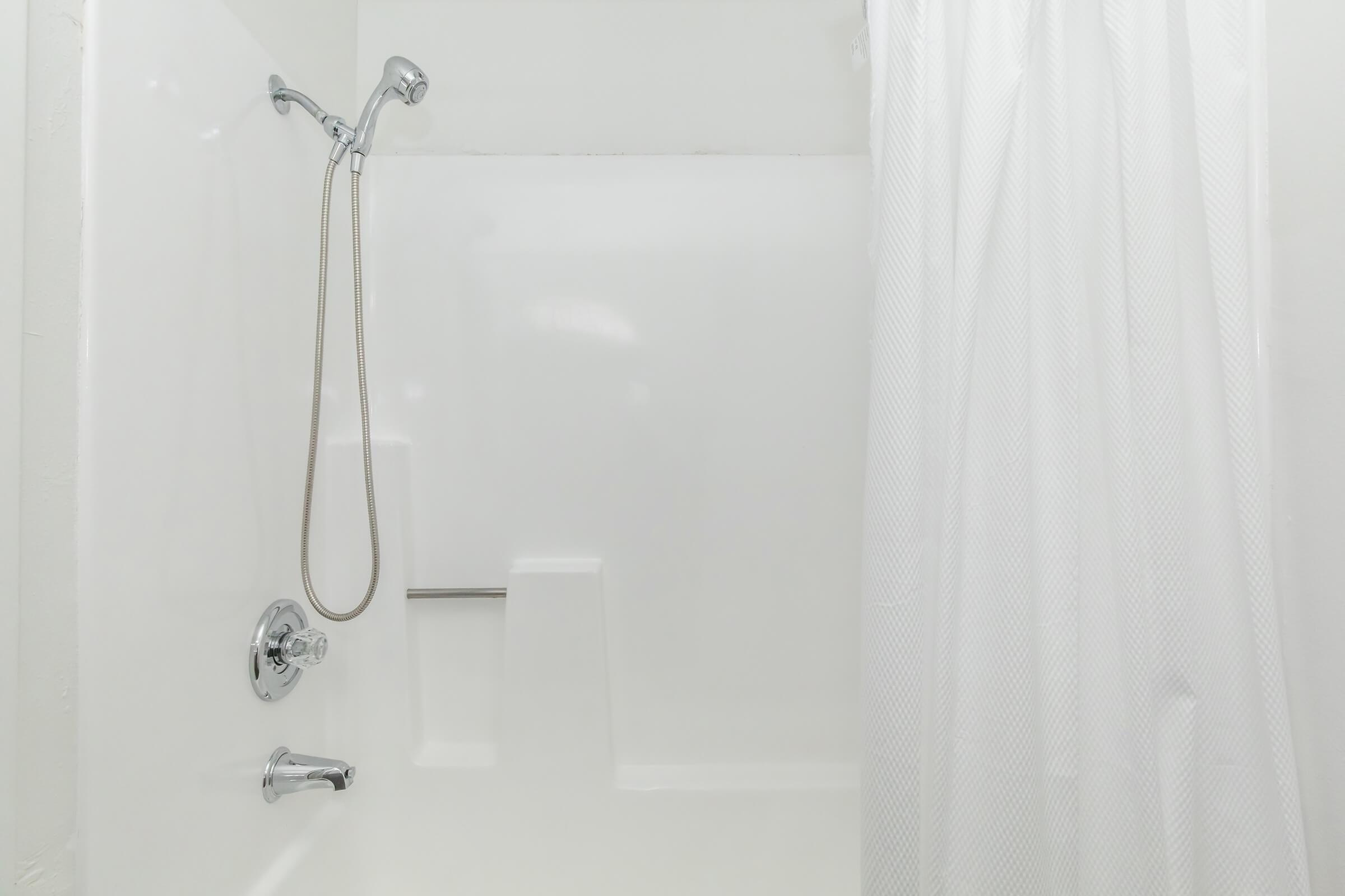 A clean, empty bathroom shower stall featuring a white shower curtain, a chrome showerhead, and a chrome faucet. The walls and floor are plain white, highlighting the minimalist design of the space.