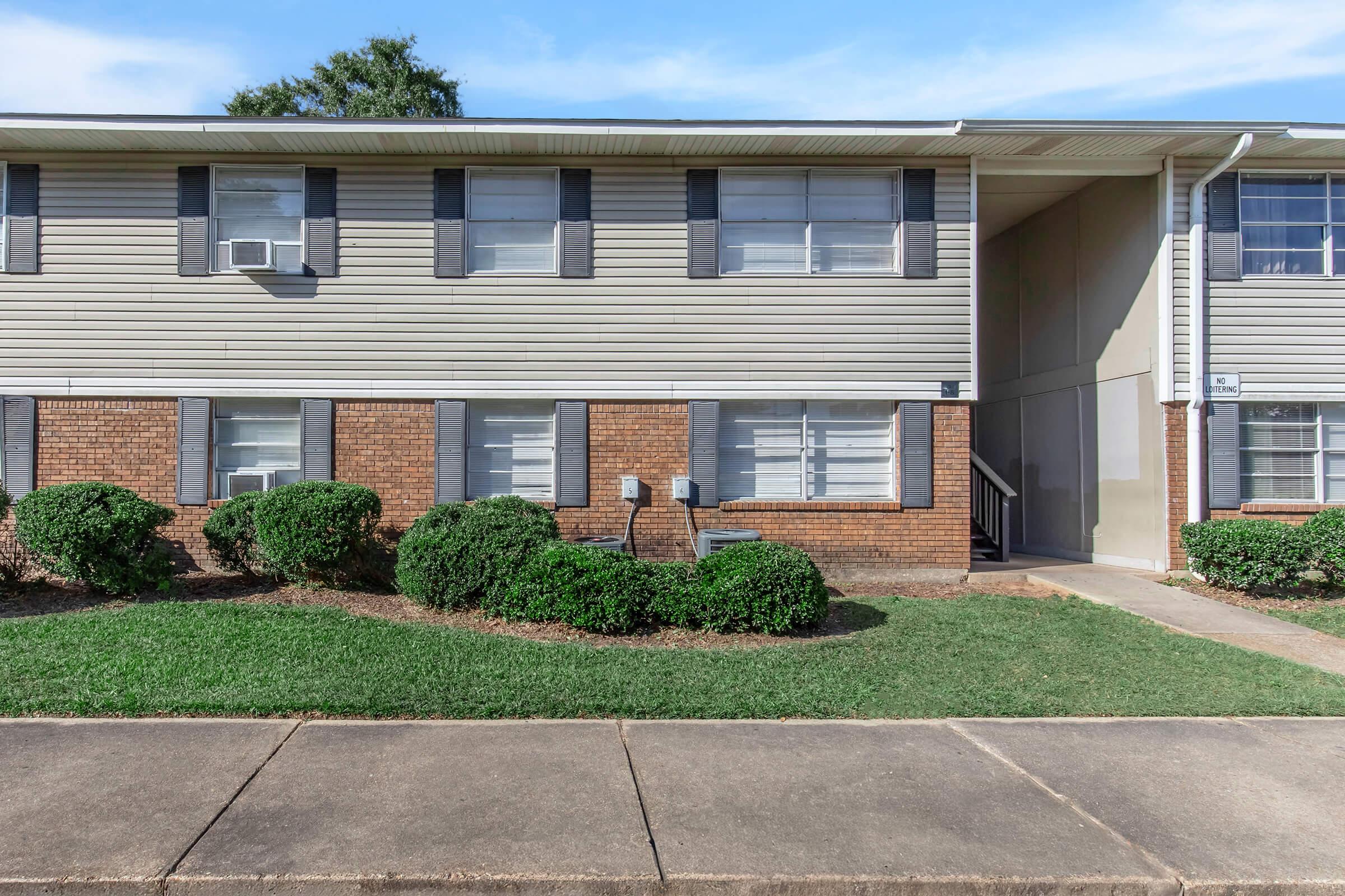 A two-story residential building with a light gray exterior and brown brick accents. The ground level features several windows, air conditioning units, and small shrubs. A concrete pathway leads up to the entrance, surrounded by grass and landscaping. The sky is clear, indicating a sunny day.