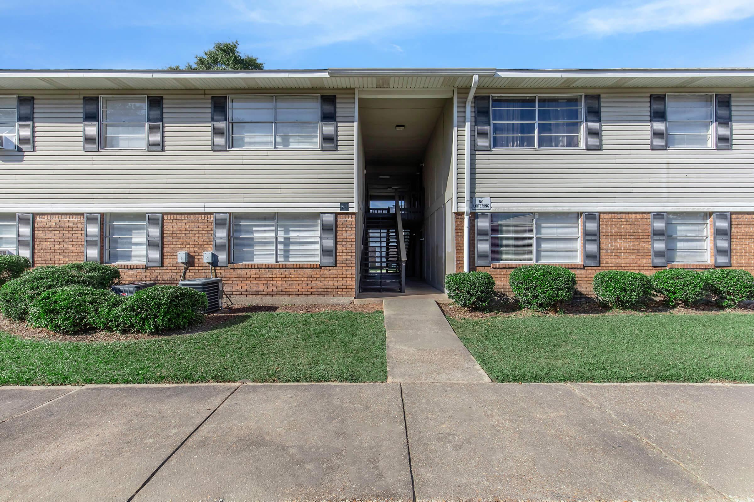 A two-story apartment building with a beige exterior and dark shutters. The entrance features an exposed staircase leading to the second floor. Surrounding the building are well-maintained shrubs and a grassy area. The pavement in front leads to the entrance, and the sky is clear with few clouds.