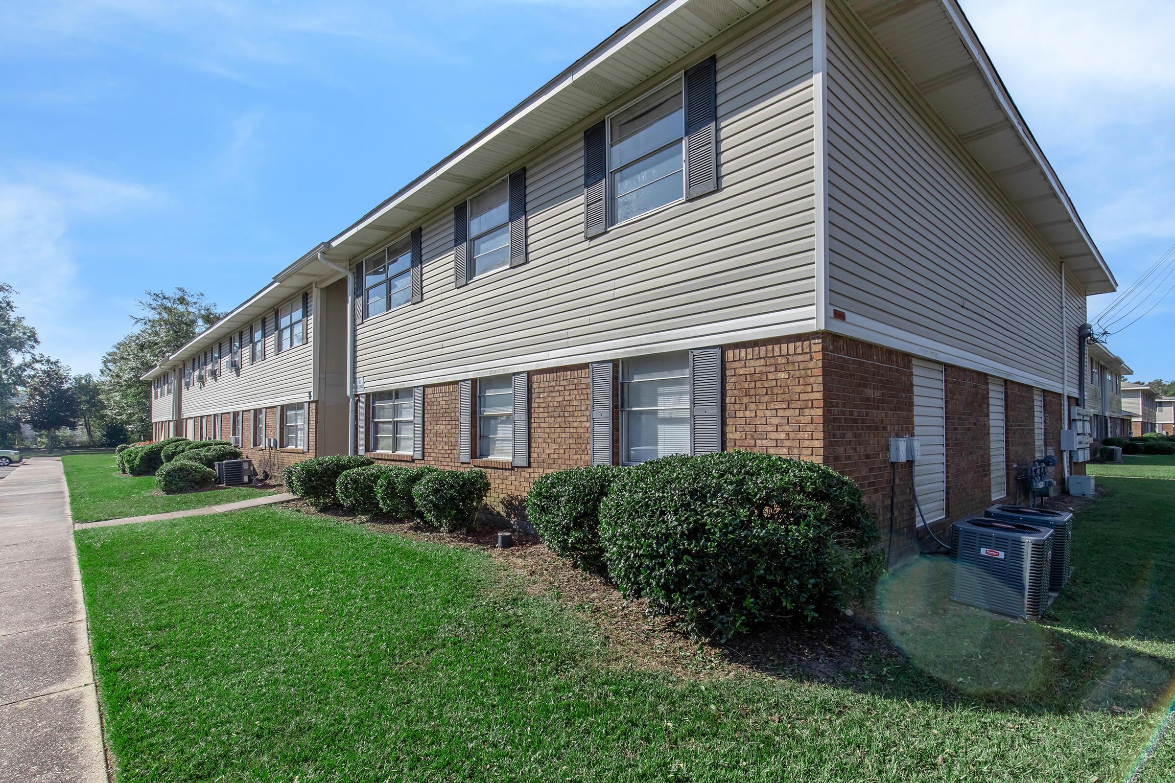 Two-story apartment building featuring a combination of brick and siding exteriors. The building is surrounded by well-manicured grass and small bushes. Sidewalks are present, leading to the entrance, with air conditioning units visible near the ground level. Bright blue sky in the background.