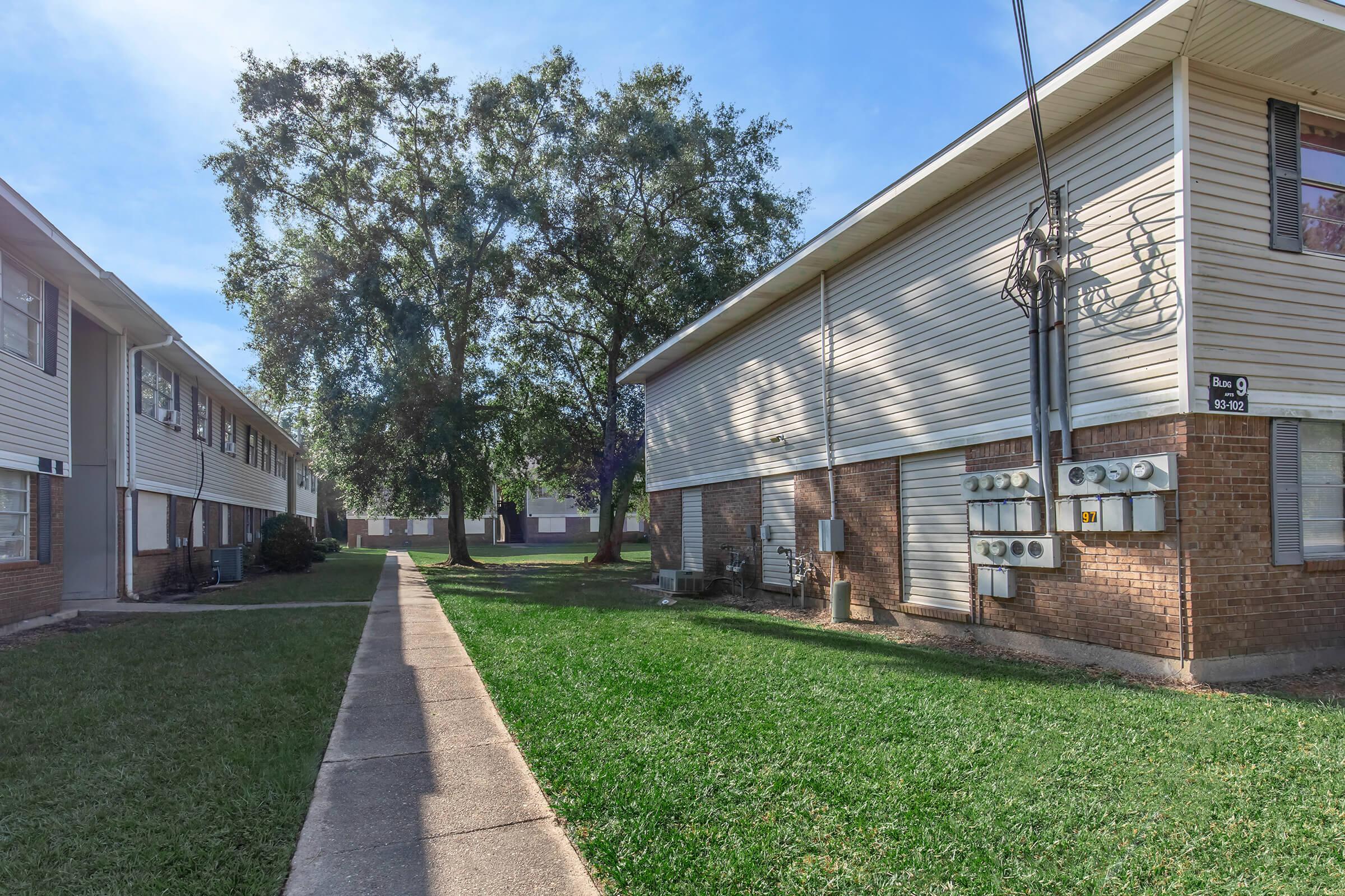A view of a residential area featuring two-story apartment buildings with electrical meters on the side of one building. There is a paved walking path lined with grass, and a few trees in the background under a clear blue sky. The setting appears calm and well-maintained.