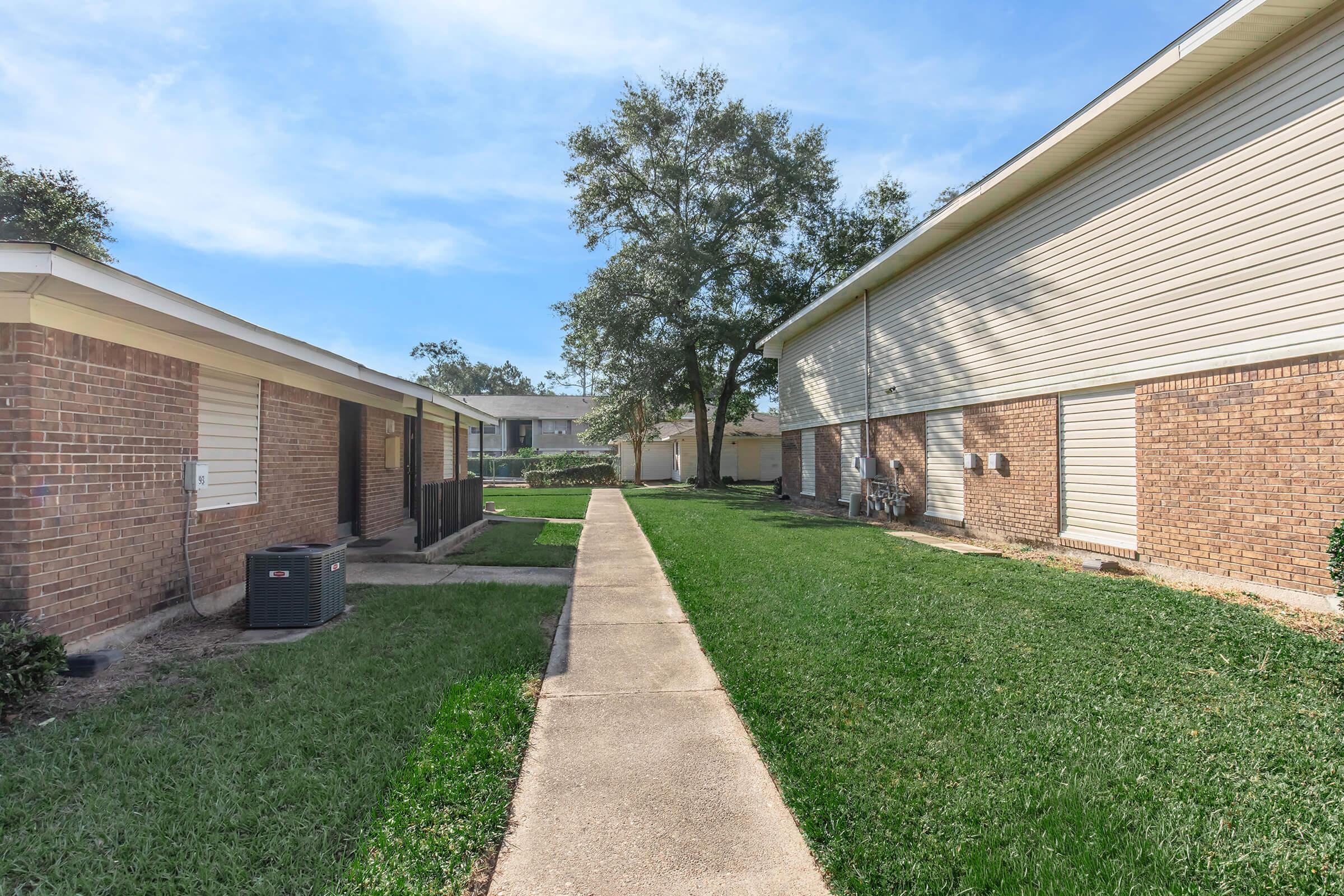 A clear view of a pathway lined with grass, leading between two brick buildings with light-colored siding. Trees are visible in the background, and there are air conditioning units along the sides of the buildings. The scene is bright and sunny, showcasing a well-maintained outdoor area.