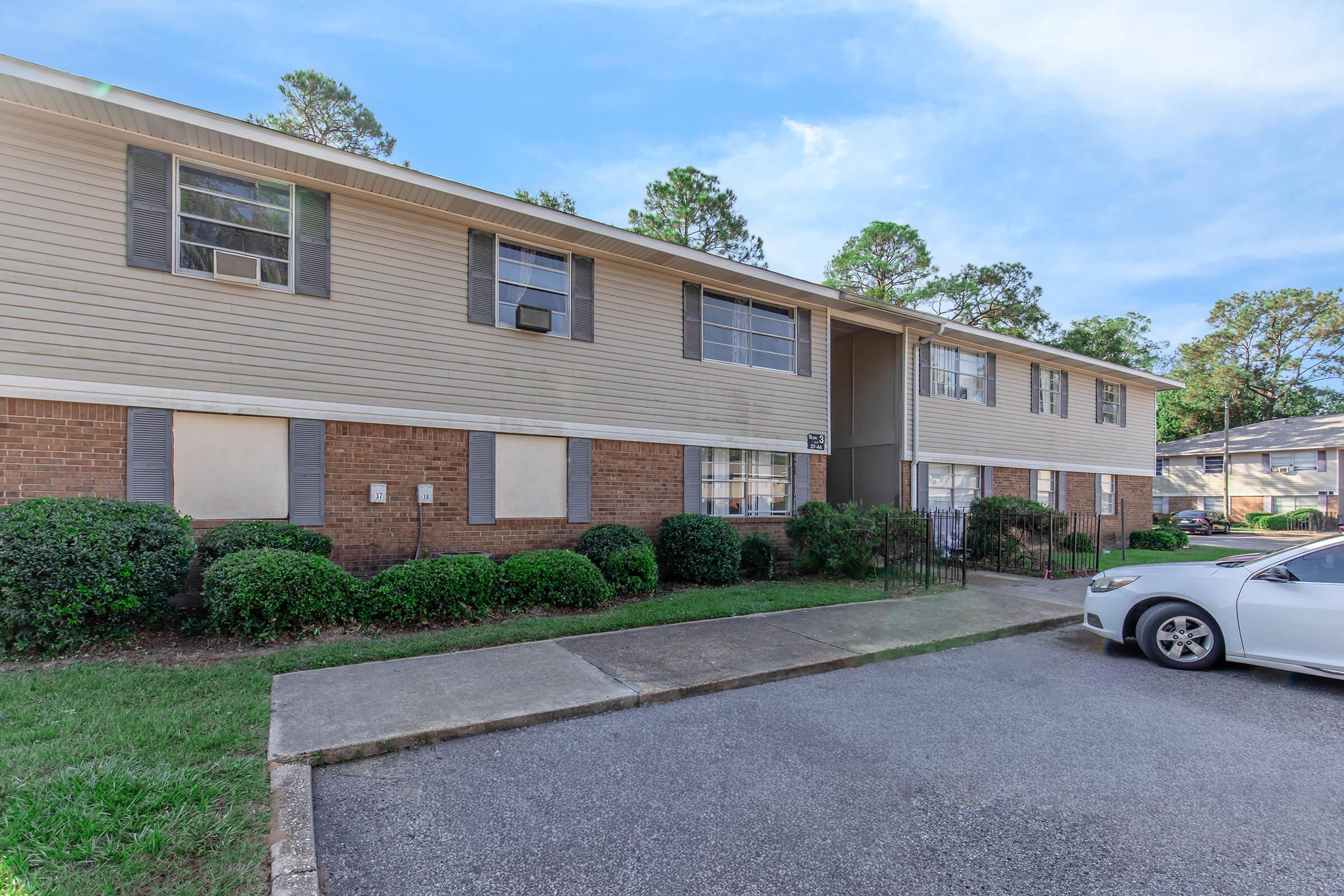 Exterior view of a two-story apartment building featuring multiple windows and a brick facade, surrounded by well-maintained shrubs and grass. A parking area is visible in front, with a white car parked nearby. The setting includes trees in the background and a clear blue sky.