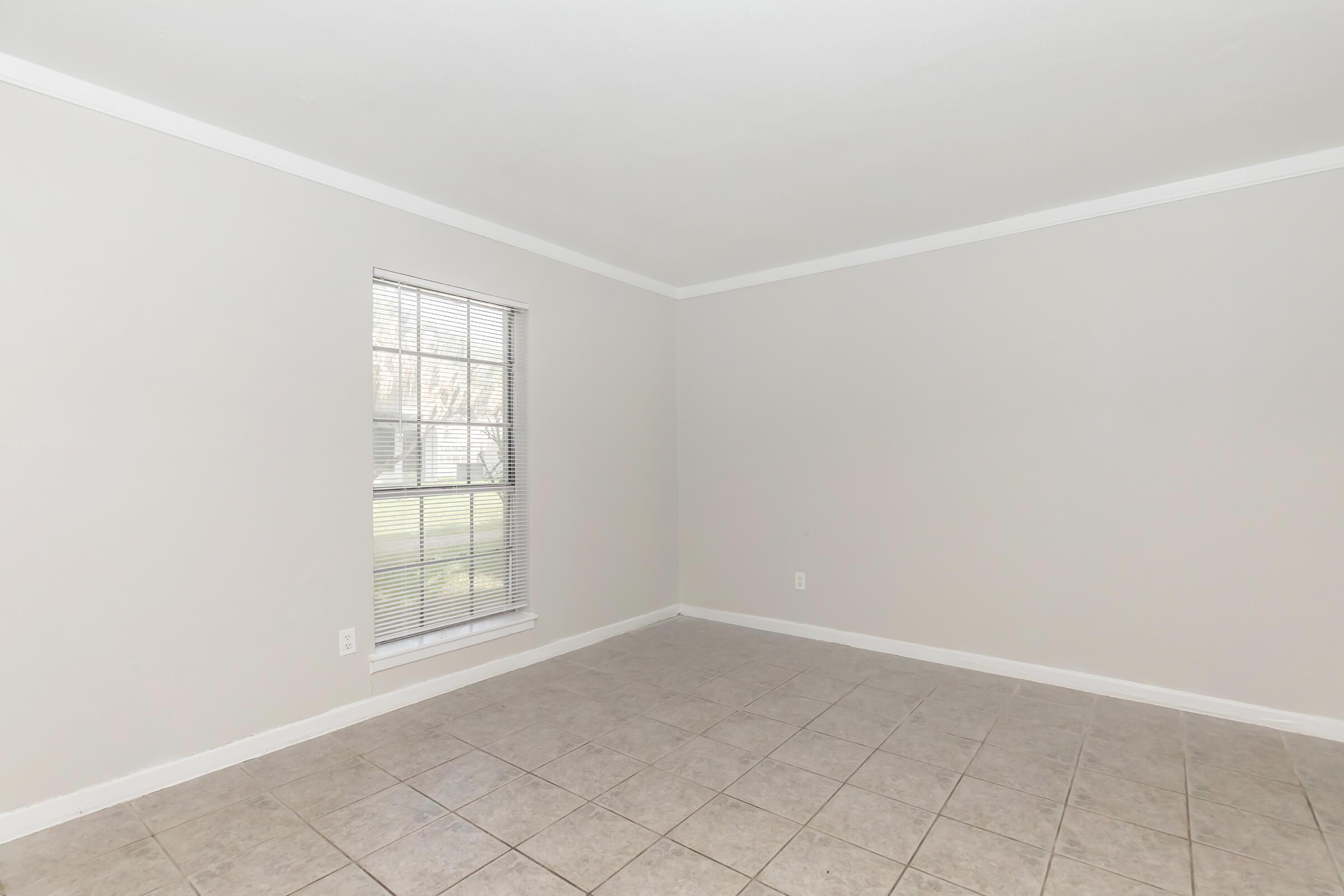Empty room with light gray walls and a window on the left side. The floor is covered with light-colored tiles. The window has white blinds partially open, allowing natural light to enter. The room is sparsely furnished, giving a spacious and clean appearance.