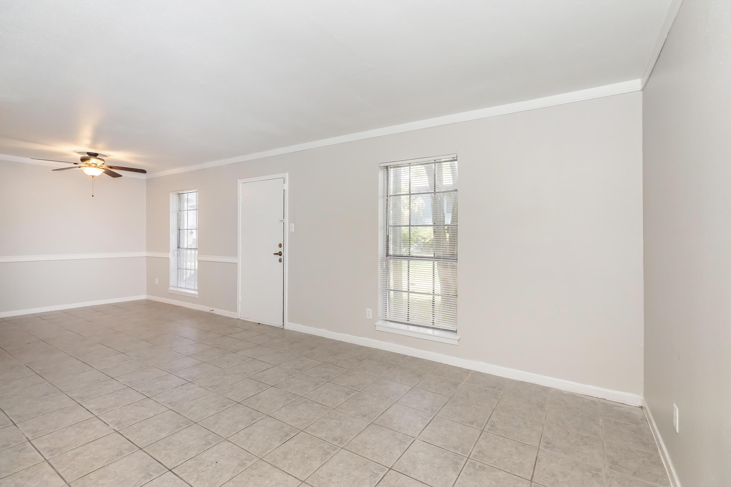 A spacious, light-colored living room featuring tiled flooring, two windows allowing natural light, and a door leading outside. The ceiling has a ceiling fan, and the walls are painted in a neutral shade, providing a bright and airy atmosphere.