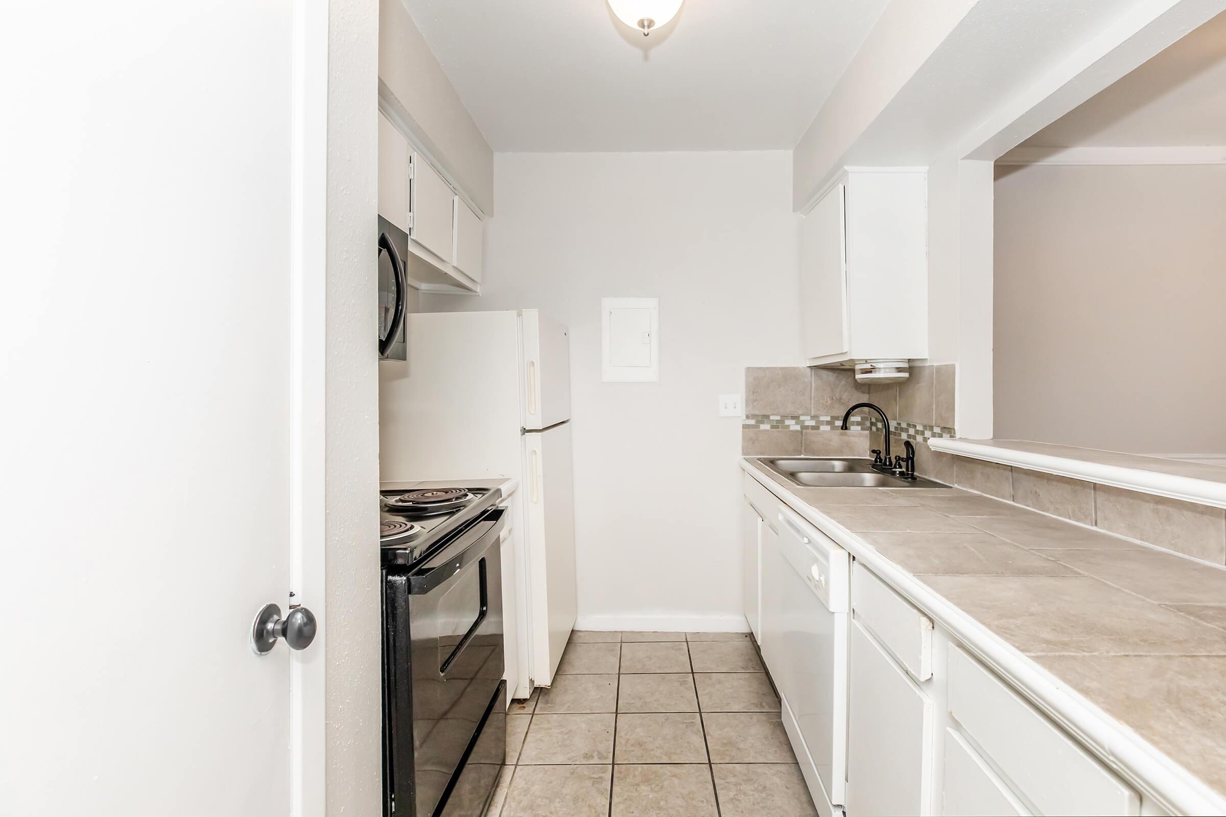 A clean, modern kitchen featuring white cabinets, a stainless steel sink, and a tile backsplash. The kitchen includes a stove, microwave, and refrigerator. The countertop is spacious, and the floor is tiled. Natural light comes from a ceiling fixture, creating a bright atmosphere.