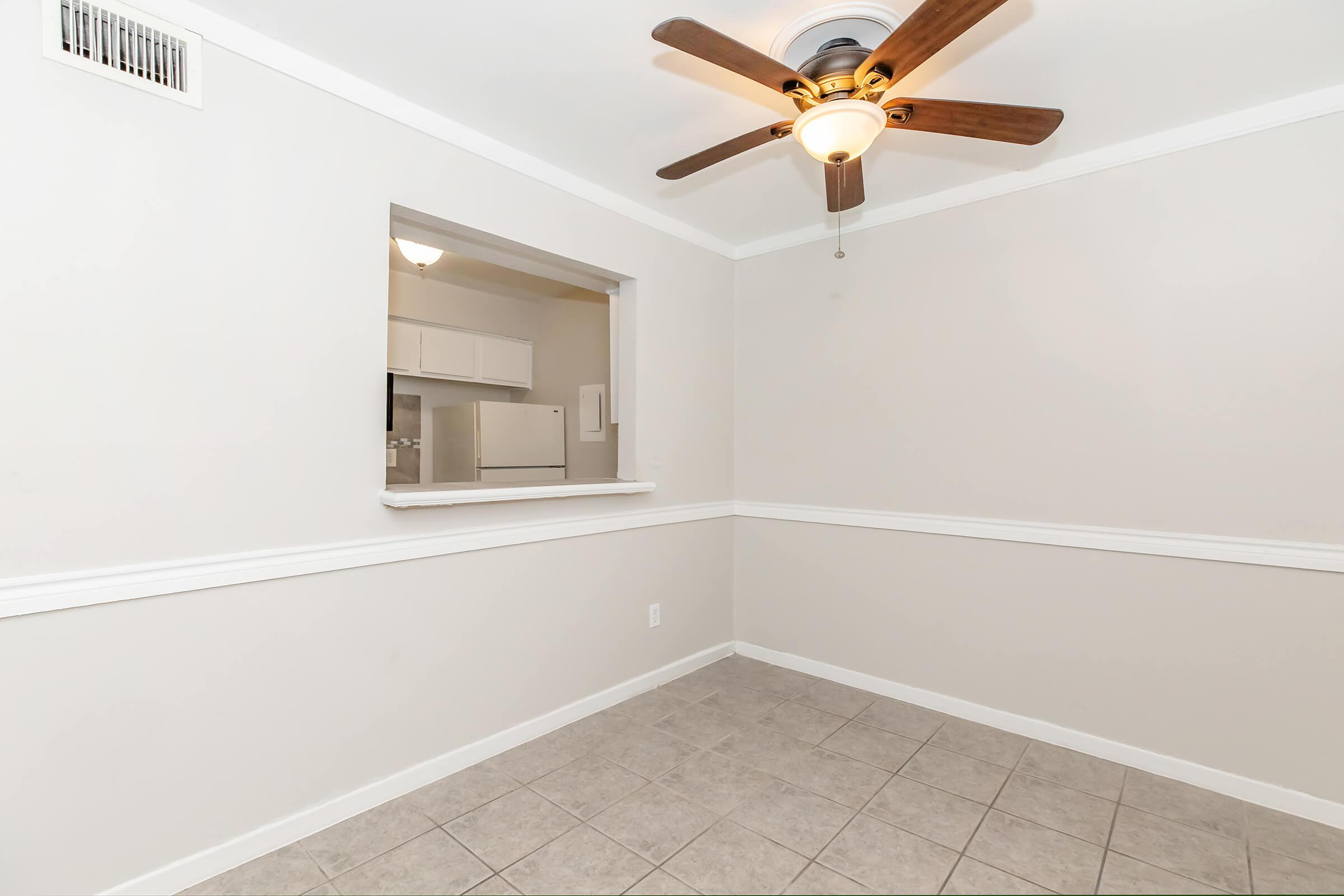 A spacious room featuring light-colored walls, a ceiling fan with wooden blades, and tile flooring. There is a wall opening that connects to a kitchen area, with a glimpse of white cabinetry and appliances visible. The room has a clean, modern look, ideal for various uses.