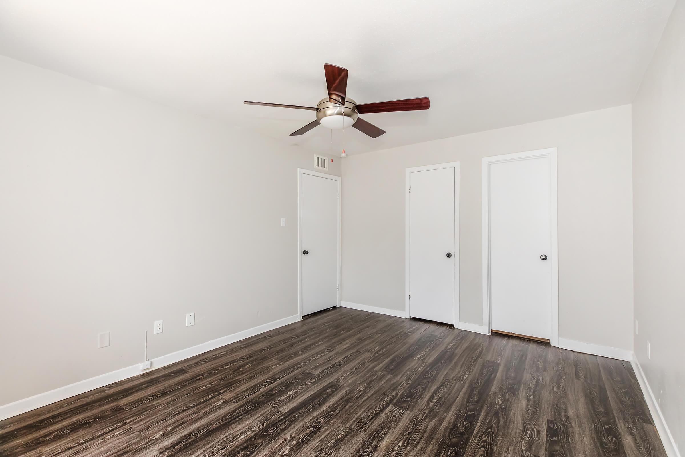 A spacious, empty room with light-colored walls and dark wooden flooring. A ceiling fan with a light fixture is mounted in the center. There are two closed white doors on the right side, and the room is well-lit from natural light. The space appears clean and ready for furnishings.