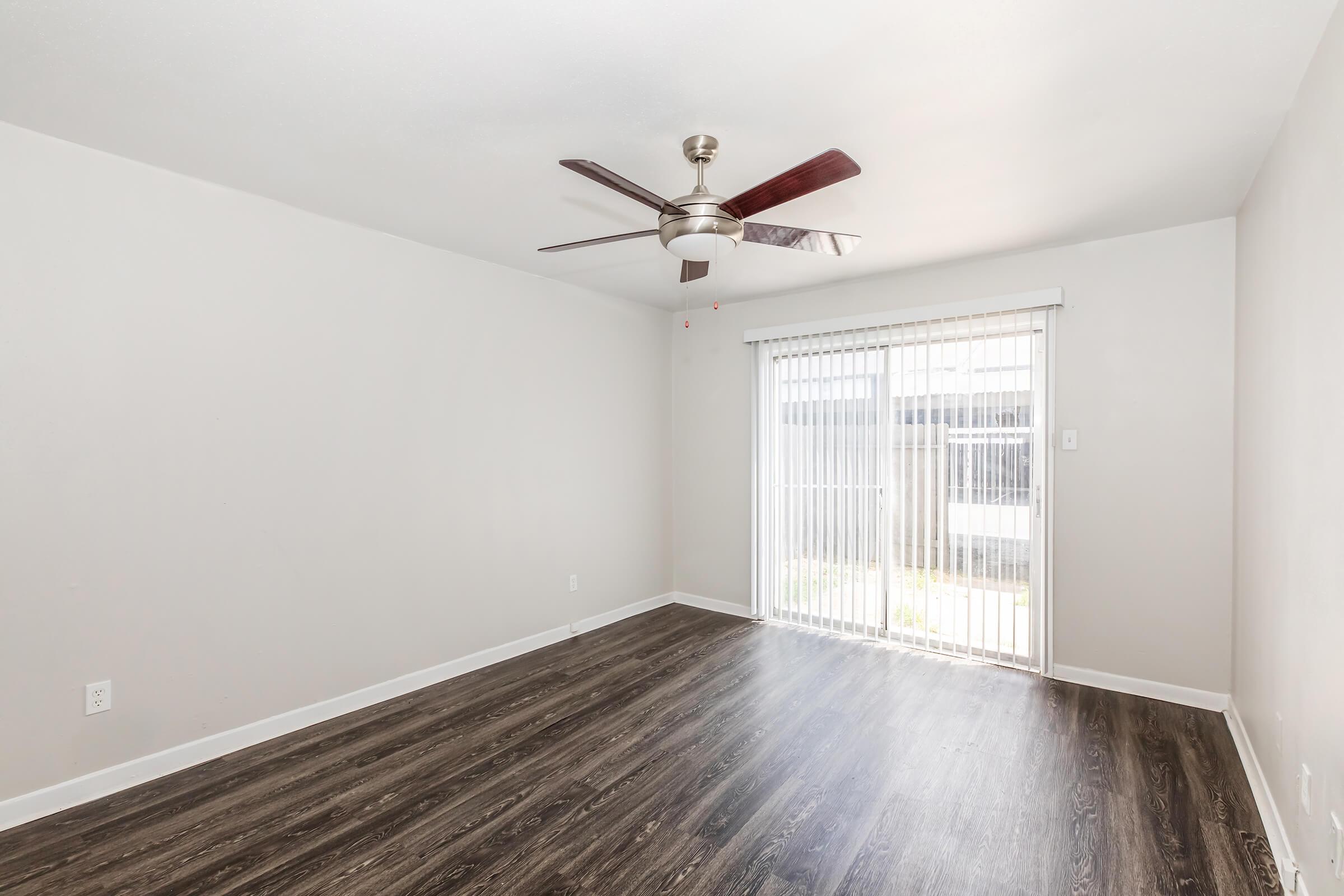 A bright, empty room with light-colored walls and dark wood-like flooring. A ceiling fan is mounted in the center, and a large window with vertical blinds allows natural light to enter. The room appears spacious and ready for furnishings.