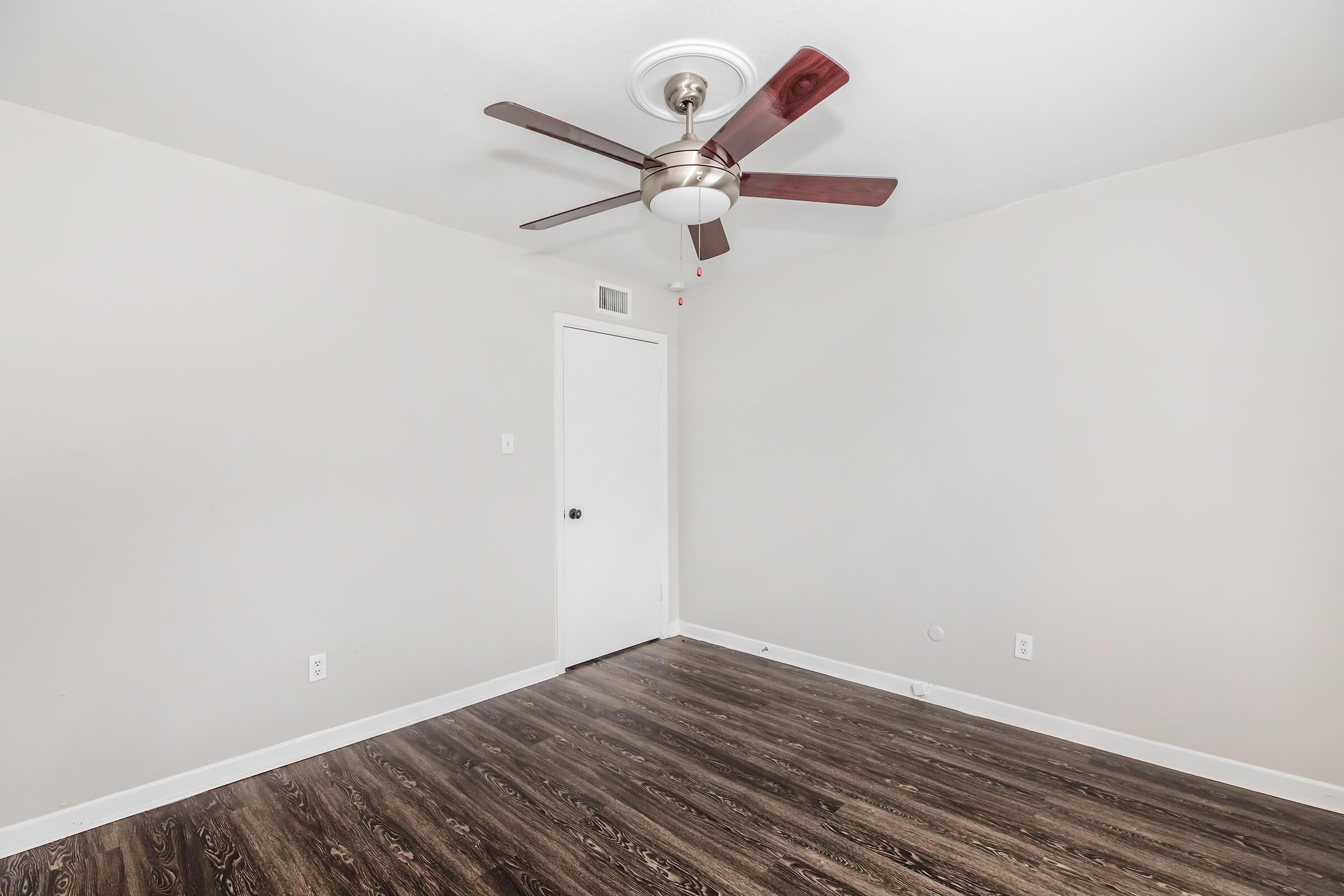 A bare room with light gray walls and dark wood-like flooring. A ceiling fan with five blades hangs in the center, and a closed white door is visible on the left. The space is empty, creating a clean and open feel.