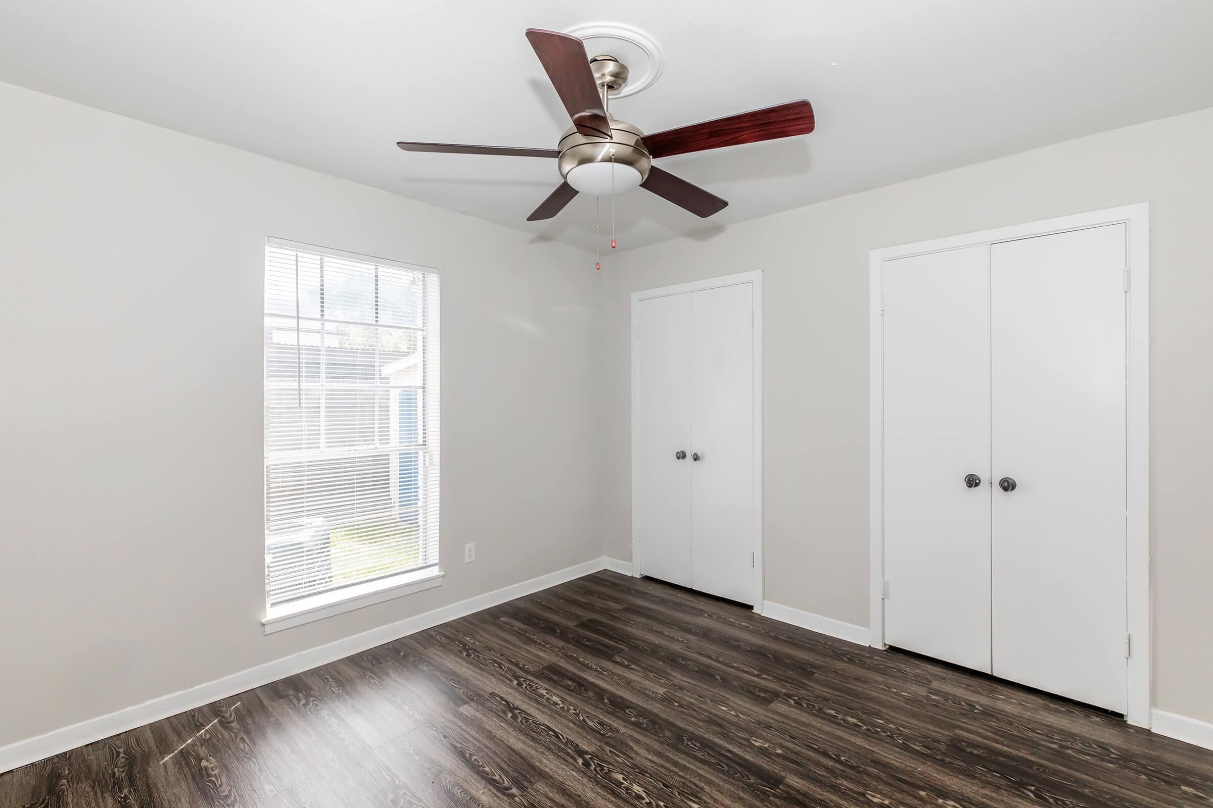 A well-lit room featuring a ceiling fan, light gray walls, and a window with blinds. The floor is dark wood-like laminate, and there are two white double-door closets on the right side. Natural light streams in through the window, creating a bright and airy atmosphere.