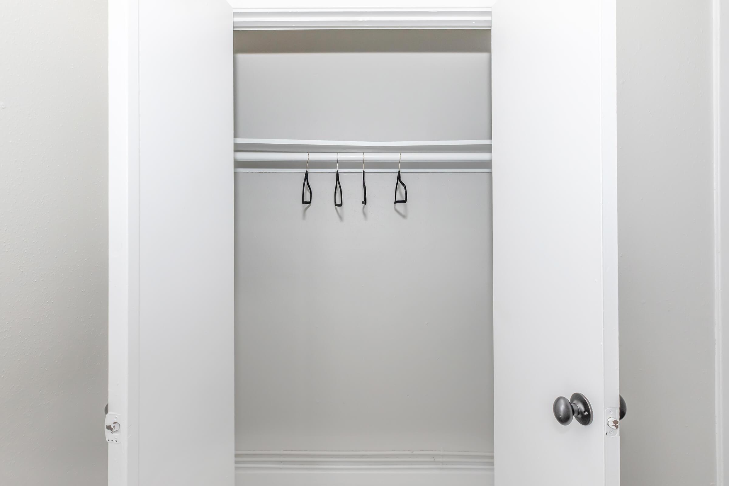 Empty closet with white walls and two open doors, featuring two horizontal hanging rods. Four black hangers are suspended from the upper rod, while the lower rod remains bare. The overall look is minimalist and organized.
