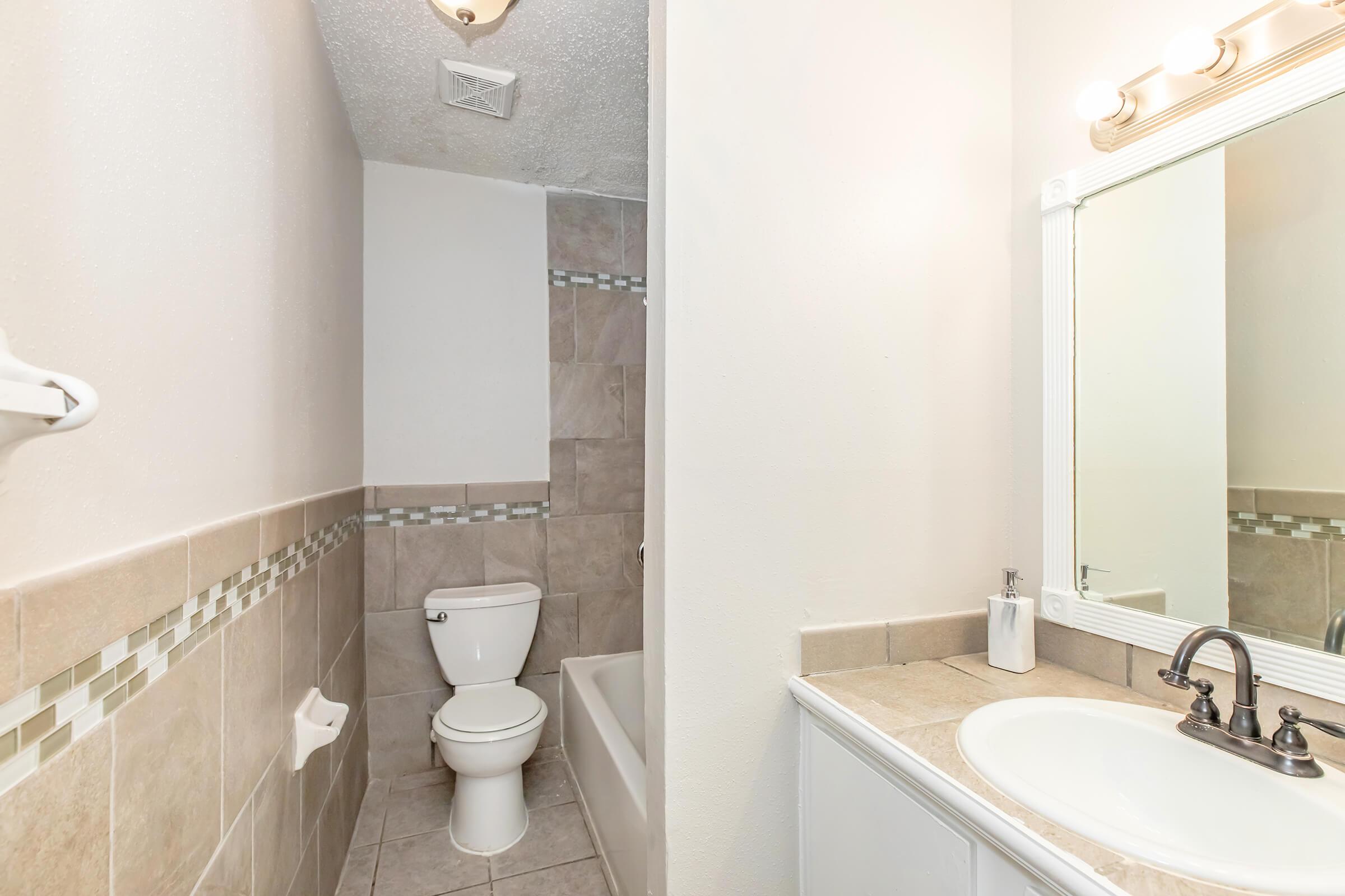 A clean bathroom featuring a white toilet, a bathtub with a neutral-colored tiled wall, and a sink with a mirror above. The flooring and wall tile incorporate a mix of beige and gray tones, adding a modern touch. Soft lighting illuminates the space.
