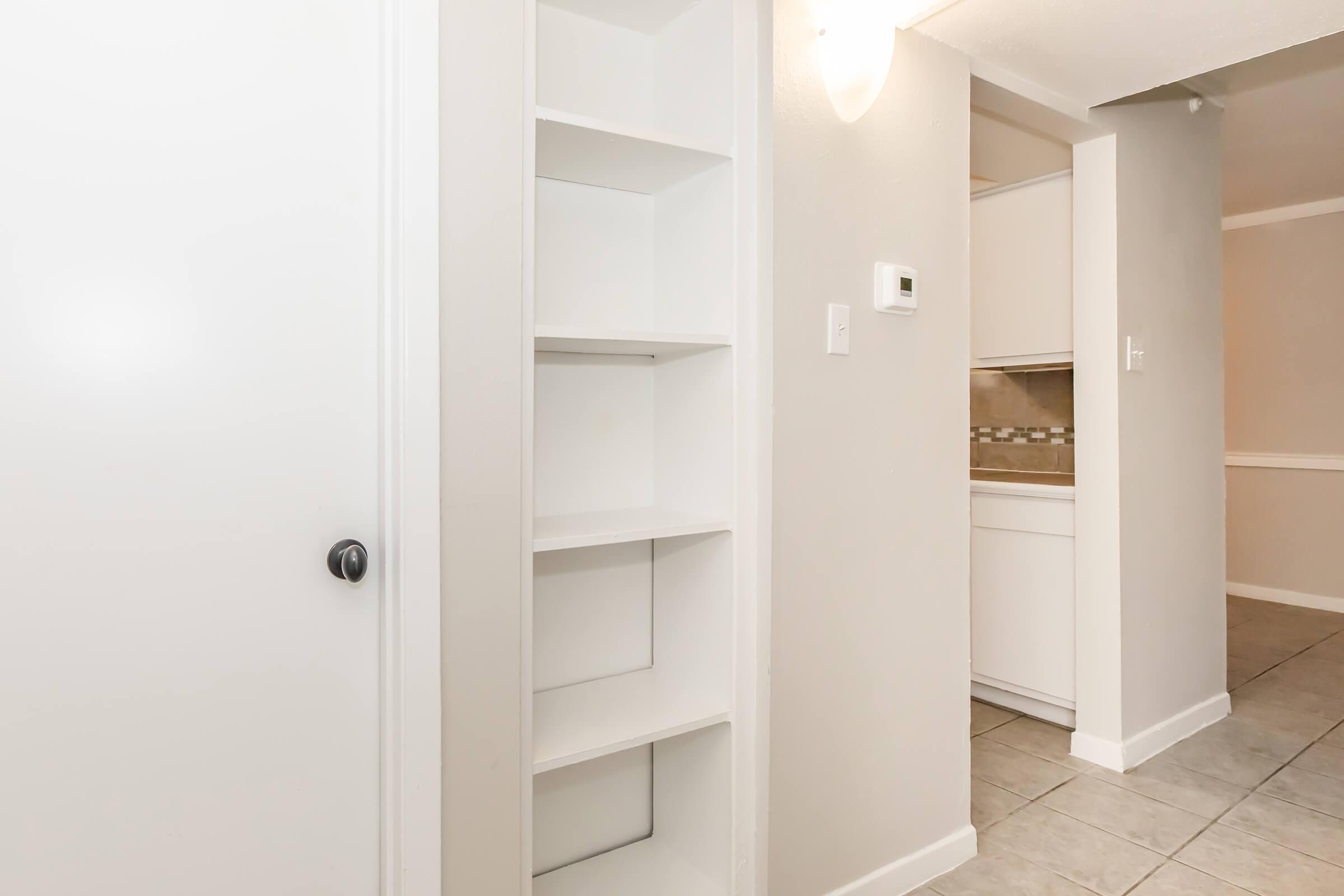 Interior view of a hallway featuring white shelving built into the wall, with a door leading to another room. The walls are painted in a light color, and there are tiled floors. A narrow opening leads to a kitchen area in the background, showcasing a clean and modern design.
