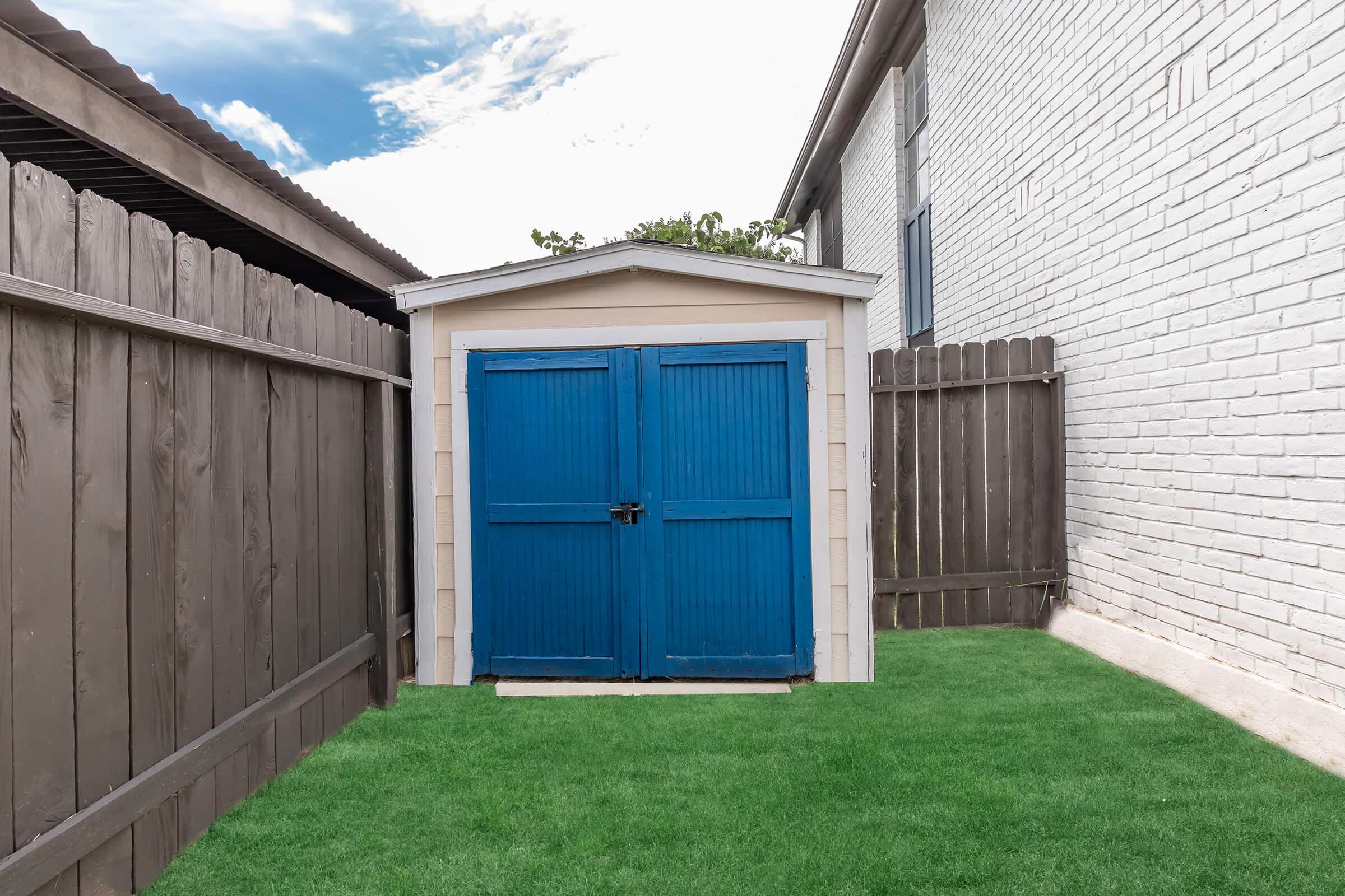 A small blue shed with double doors sits in a backyard, surrounded by wooden fences. The shed is situated on a patch of green grass, with a clear sky and scattered clouds overhead. The shed's design is simple and functional, providing storage space within a residential setting.