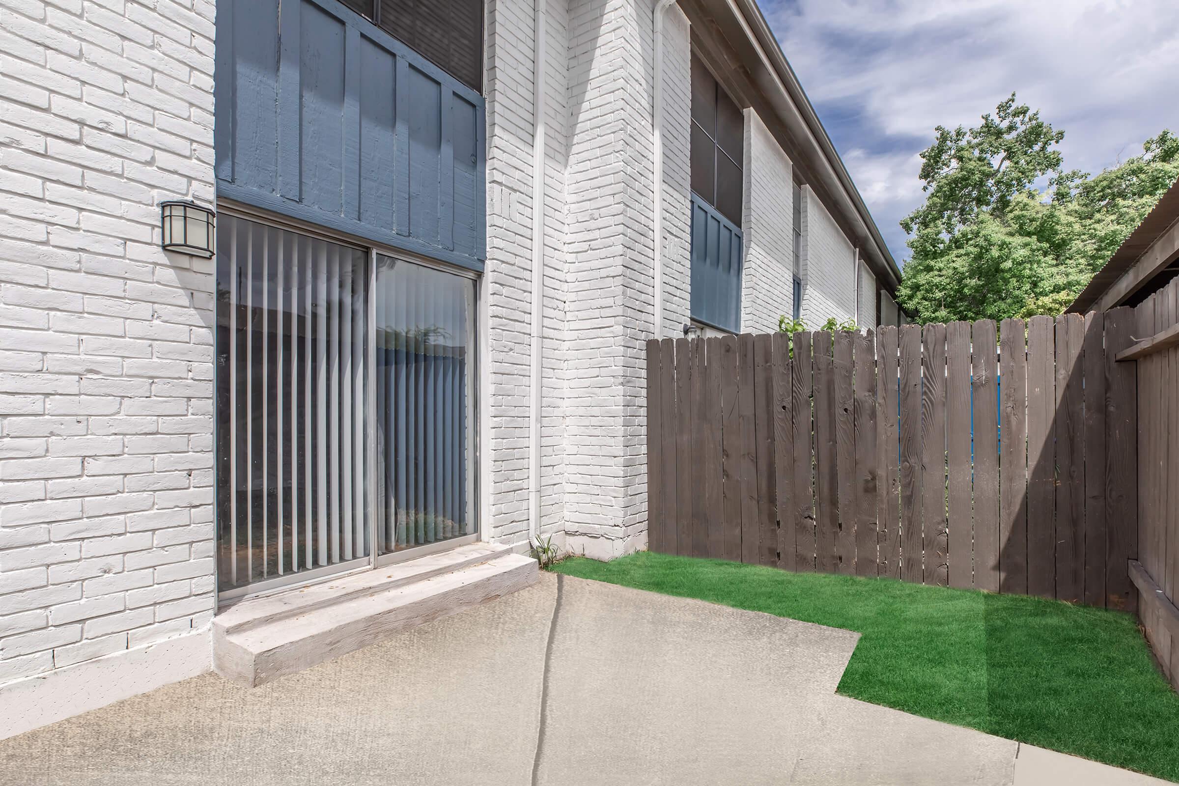 A corner of a modern outdoor area featuring a white brick wall, a sliding glass door with vertical blinds, and a wooden fence. The ground is a mix of concrete and artificial grass, with trees visible in the background under a partly cloudy sky.